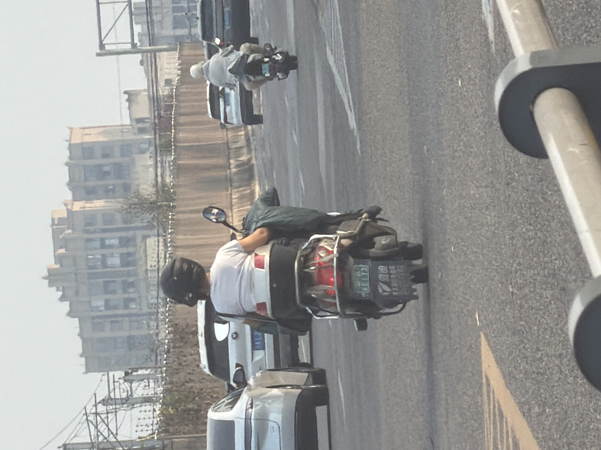 In the city of Jiangjin, China, a motorcycle is traveling on a road. The rider, wearing a black helmet and a white t-shirt, is in the foreground. Behind the motorcycle, a white SUV is visible. In the distance, a large, multi-story building with a distinctive architectural style stands out against the hazy sky. The road appears to be a highway or a major thoroughfare, with other vehicles and road markings indicating traffic. The overall scene depicts a typical urban environment in China during daylight hours.