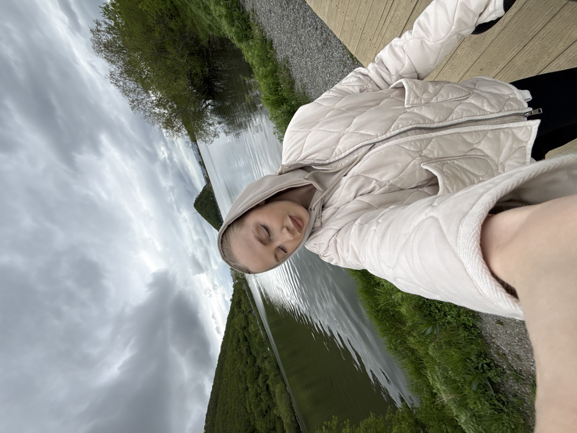 A young woman, with blonde hair pulled back, is taking a selfie outdoors by a body of water. She is wearing a light beige hooded jacket over a similar colored hoodie and black pants. Her eyes are closed, and her head is tilted down. The background features a large, calm body of water, with green, tree-covered hills on the left and right sides. The sky is overcast with dark, dramatic clouds. A portion of a wooden deck is visible in the foreground on the right. The location is the Staraya Pochtovaya recreation area in Petropavlovsk-Kamchatsky, Russia, a popular spot for locals with views of the surrounding natural landscape.