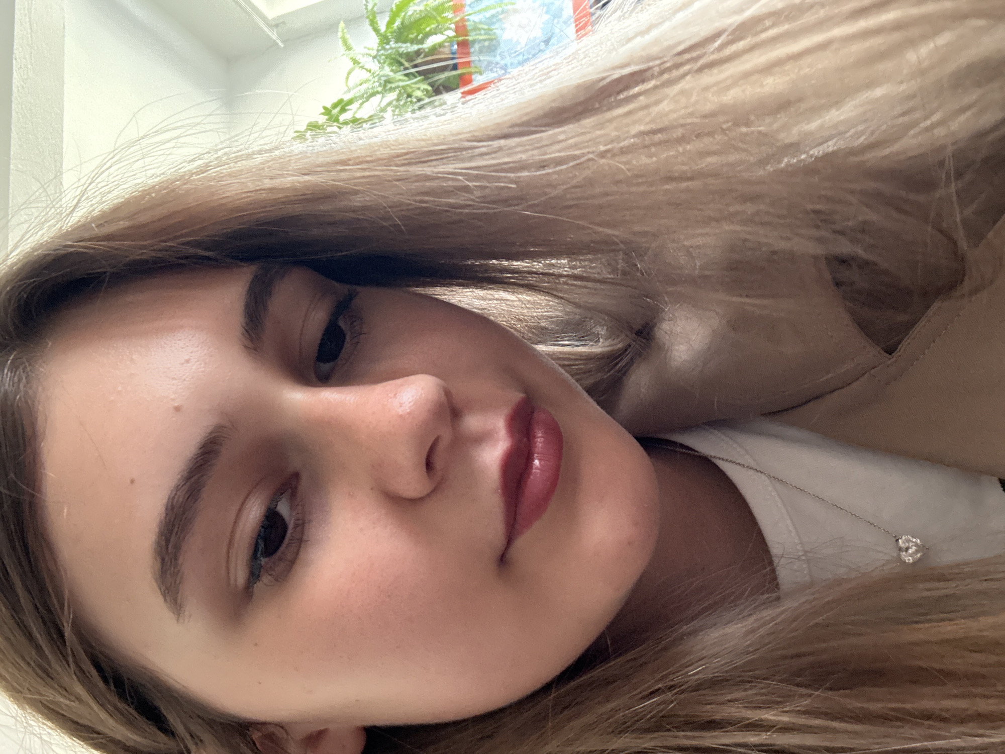 A close-up, rotated image shows a young woman with fair skin and light brown hair. She has dark brown eyes, a small mole on her forehead, and full lips adorned with a glossy, plum-colored lipstick. She is wearing a white top and a light beige jacket. A silver necklace with a small heart pendant is visible against the white top. In the background, partially out of focus, there is a window with a green plant. The location in Panagyurishte, Bulgaria, cannot be determined from the visual information provided.