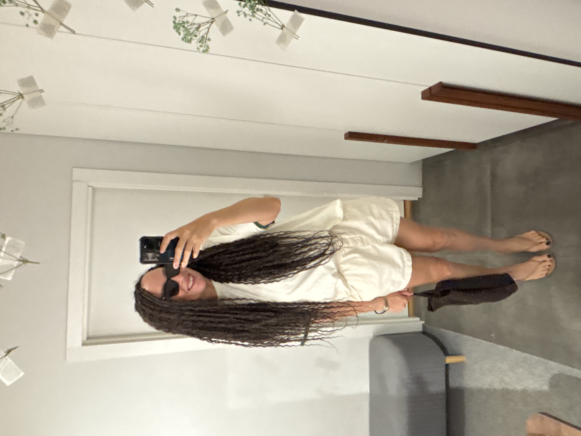 The image depicts a woman taking a mirror selfie indoors in Chaikovsky, Russia. The woman, who appears to be in her 20s or 30s, has long, dark, micro-braided hair and is smiling, with her teeth visible, while wearing dark sunglasses. She holds a black smartphone in her right hand, positioned to capture her reflection. She is dressed in a loose-fitting, cream-colored short-sleeved t-shirt with a green stripe on the left sleeve cuff and matching cream-colored shorts with an elastic waistband. A watch with a brown strap and a blue bracelet is visible on her left wrist. She carries a small, dark brown or black woven bag in her left hand. Her bare legs are visible, and she wears light-colored flip-flops with black-painted toenails. The indoor environment features white walls and a dark grey floor. A grey upholstered bench with light wooden legs is situated to the left. The mirror on the right reflects the scene and reveals small, dried green botanical decorations taped to the wall with white adhesive strips, arranged vertically. Two light-colored, geometric decorative fixtures hang from the ceiling above. A white doorframe is partially visible in the background to the left.