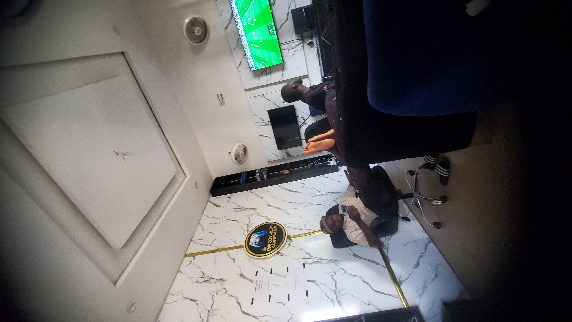 This image depicts the interior of King Zion Luxury Games Palace, located in Jalingo, Nigeria. The establishment features walls adorned with white marble-patterned tiles, accented by vertical gold trim. The ceiling is a multi-level white drop design with recessed lighting fixtures and two visible circular ceiling fans. The floor is covered with light brown tiles.
Three adult males are present within the space. On the left, one male is seated in a black office-style chair with wheels, wearing a light-colored short-sleeved collared shirt, dark trousers, and a baseball cap. He is holding and viewing a smartphone. In the center-right, another male is reclining on a dark sofa, with his bare feet propped up. He is wearing dark trousers and a dark top, and his head is turned towards a television screen. A third male is seated further to the right, also facing a television screen, wearing a dark shirt.
A prominent circular sign on the left wall displays "KING ZION LUXURY GAMES PALACE" with a white game controller graphic. Multiple black flat-screen televisions are mounted on the walls. The largest television on the right displays a soccer video game interface. A tall, narrow black shelving unit is positioned against the wall to the right of the main sign. A pair of black Adidas slides with white stripes are visible on the floor near the feet of the male reclining on the sofa. Several small, white handwritten or printed notices are affixed to the left marble wall.