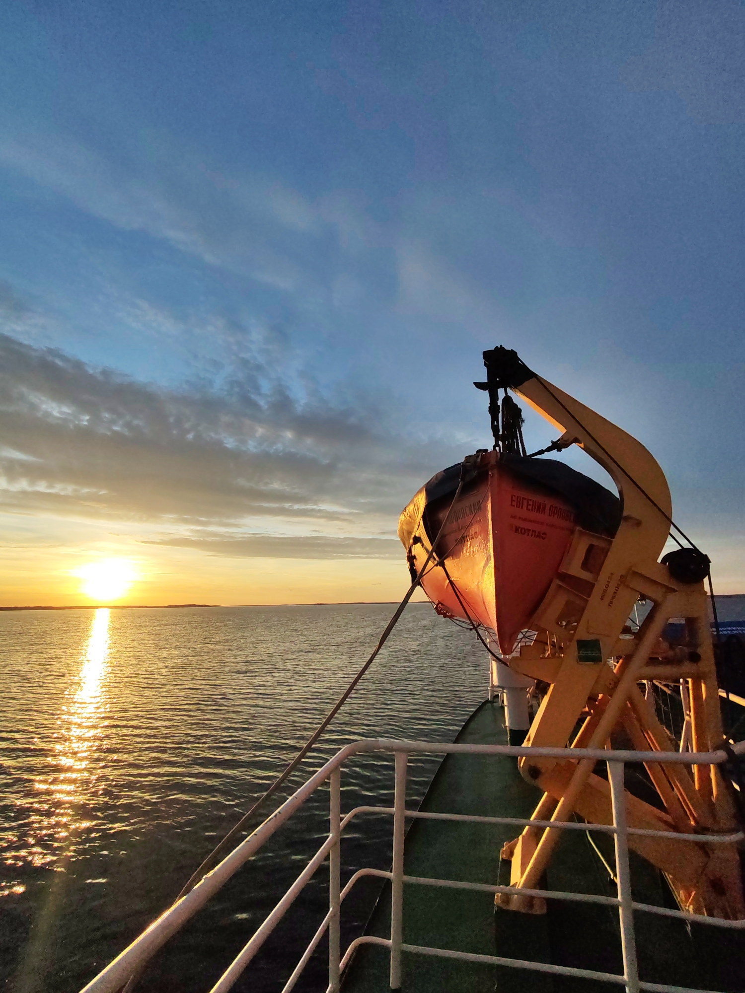 The image shows a lifeboat secured on a davit on a ship at sunset. The lifeboat is orange and has Russian writing on it, "Евгений Орловский Котлас" (Evgeniy Orlovskiy Kotlas), indicating a potential location near Kotlas, Russia, or a vessel associated with the city. The davit is yellow. The sun is setting over the water, reflecting light onto the sea surface. The sky has a gradient from blue to orange with some clouds. The ship's railing is visible in the foreground.