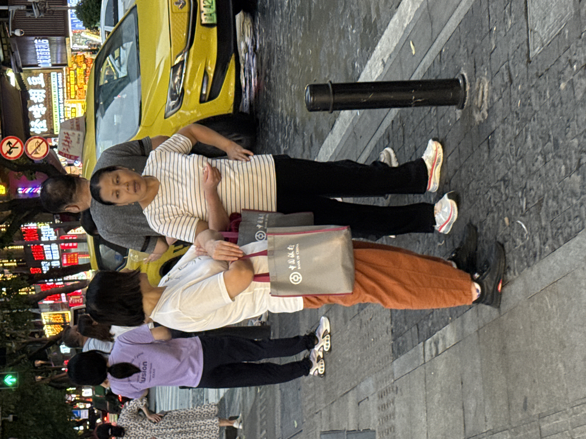 In Yuzhong District, China, at street level, a group of individuals stand on a paved sidewalk adjacent to a street where a yellow taxi is present. Two women are prominently featured. One woman, wearing an orange skirt and a white t-shirt, holds two gray shopping bags, one with "BANK OF CHINA" visible. She stands with her arms crossed. Next to her, another woman in a horizontally striped t-shirt and black pants also has her arms crossed. Behind them, a man wearing a gray t-shirt stands near the yellow taxi. Further in the background, a woman in a patterned dress and a woman in a purple t-shirt with the word "fusion" are visible. The environment is illuminated by streetlights and neon signs from nearby businesses. The ground is wet, indicating recent rain or street cleaning. A black bollard is positioned on the sidewalk to the right of the two central figures.
