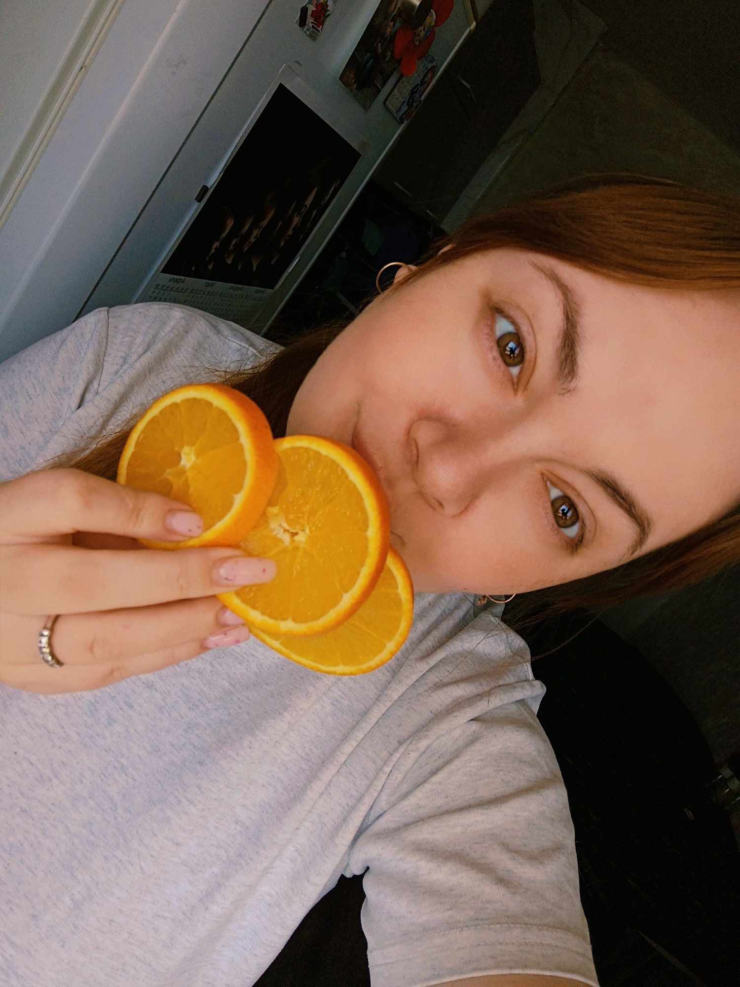 A young woman with light brown hair and hazel eyes is holding three slices of orange in her left hand close to her face in what appears to be a selfie. She is wearing a light gray t-shirt and a ring on her left ring finger. She has pierced ears with gold hoop earrings. The background includes a white door frame, a wall with what appear to be pictures or magnets on it, and a dark area. It's difficult to determine the exact location in Chaikovsky, Russia, without additional context or landmarks.