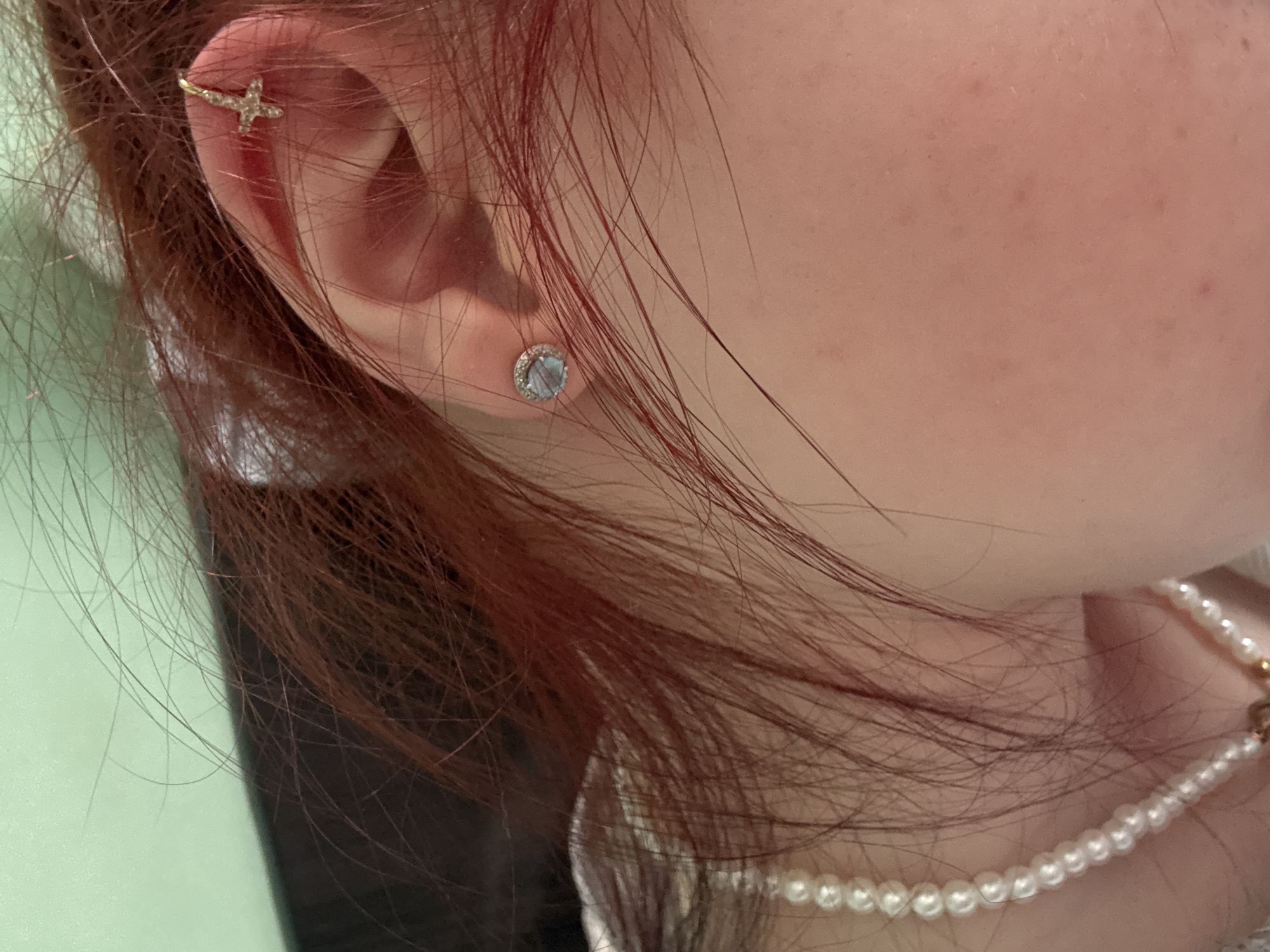A close-up, profile view shows the right ear of a person with reddish-brown hair. The earlobe has a single stud earring with a light blue stone surrounded by a halo of small, clear stones. The upper cartilage of the ear features a delicate gold earring shaped like a cross adorned with small, clear stones. A pearl necklace is visible on the person's neck. The background is a soft, pale green wall.