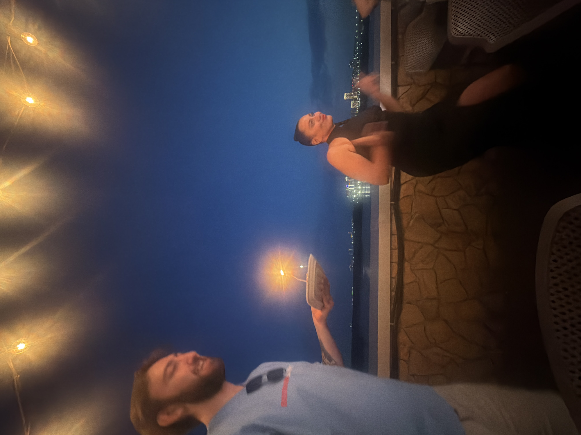 The media file depicts a group of people enjoying themselves at dusk on a balcony or patio overlooking a city skyline. The sky is a deep blue, transitioning from the lighter hues of sunset. String lights, emitting a warm yellow glow, are visible in the foreground, adding to the festive atmosphere.
Two men are the primary subjects. One, in the foreground, is laughing or smiling widely, looking upwards. He has a beard and is wearing a light blue shirt with sunglasses perched on it. The other man, further in the background, is wearing a dark top and appears to be in motion, with his arm raised as if gesturing or holding something. He has short, dark hair and is looking towards the man in the foreground.
In the background, a city skyline with numerous illuminated buildings is visible across a body of water. The architecture suggests an urban environment, and the lights indicate it is evening. The setting appears to be a relaxed outdoor gathering, possibly a party or a social event.
A notable detail is a small candle on what appears to be a piece of food, possibly a dessert, being held up by the man in the background. This suggests a celebration or a special occasion. The overall mood is joyful and convivial, with the subjects appearing to be having a good time. The time of day is clearly dusk or early evening, as indicated by the dark blue sky and the city lights beginning to shine. The weather is not explicitly discernible but the outdoor setting suggests it is pleasant.