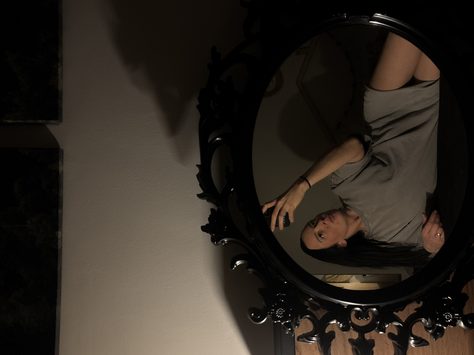 The photograph appears to be a selfie taken in a dimly lit indoor environment. The subject, a woman, is reflected in an ornate, dark-framed round mirror. She is holding a phone in her hand, presumably to take the picture. She has dark hair and is wearing a light gray, casual top. Her reflection shows part of her upper legs, suggesting she might be wearing shorts or a short skirt. The mirror is mounted on a wall, and parts of the wall are visible around the mirror, along with some decorative elements and a couple of rectangular dark frames on the wall, seemingly paintings or photos. The lighting is low, and the mirror reflects back into a room that has wood furniture.
The location is not indicated on the picture.