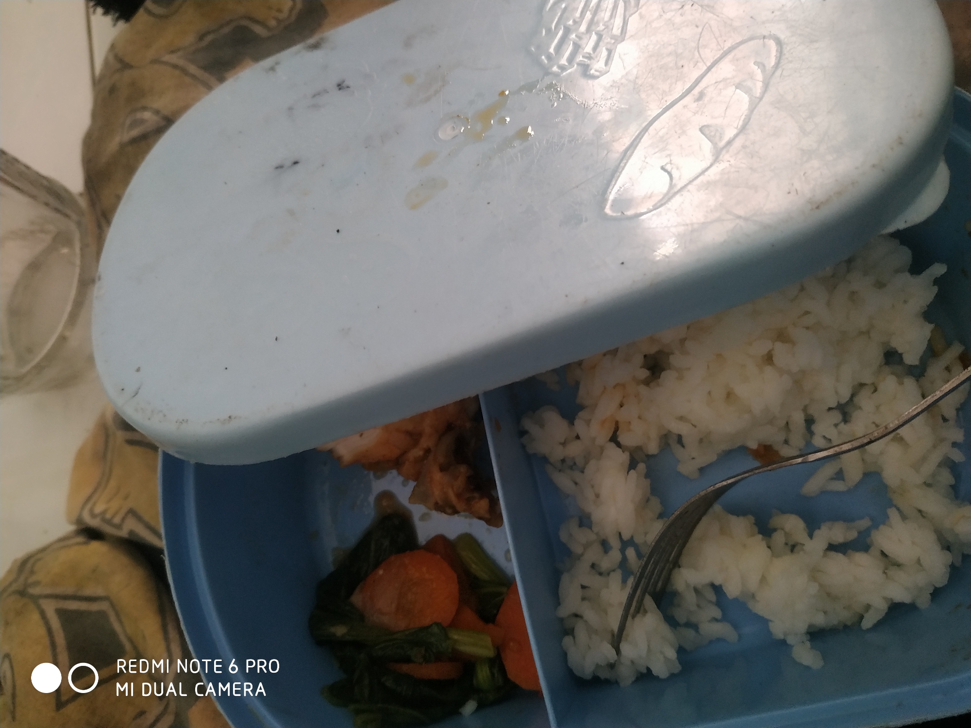 An overhead, close-up shot shows a light blue plastic bento box filled with food and a slightly ajar lid. The bento box has two compartments. The left compartment contains cooked chicken or fish pieces, green leafy vegetables, and sliced carrots. The right compartment is filled with white rice, and a silver fork is partially submerged in it. The lid is light blue and appears to have some residue or smudges on its surface. The background shows a blurred image of what appears to be a patterned textile or rug. The text "REDMI NOTE 6 PRO MI DUAL CAMERA" is visible in the lower-left corner, indicating the device used to capture the image. The scene is indoors, likely on a table or flat surface.