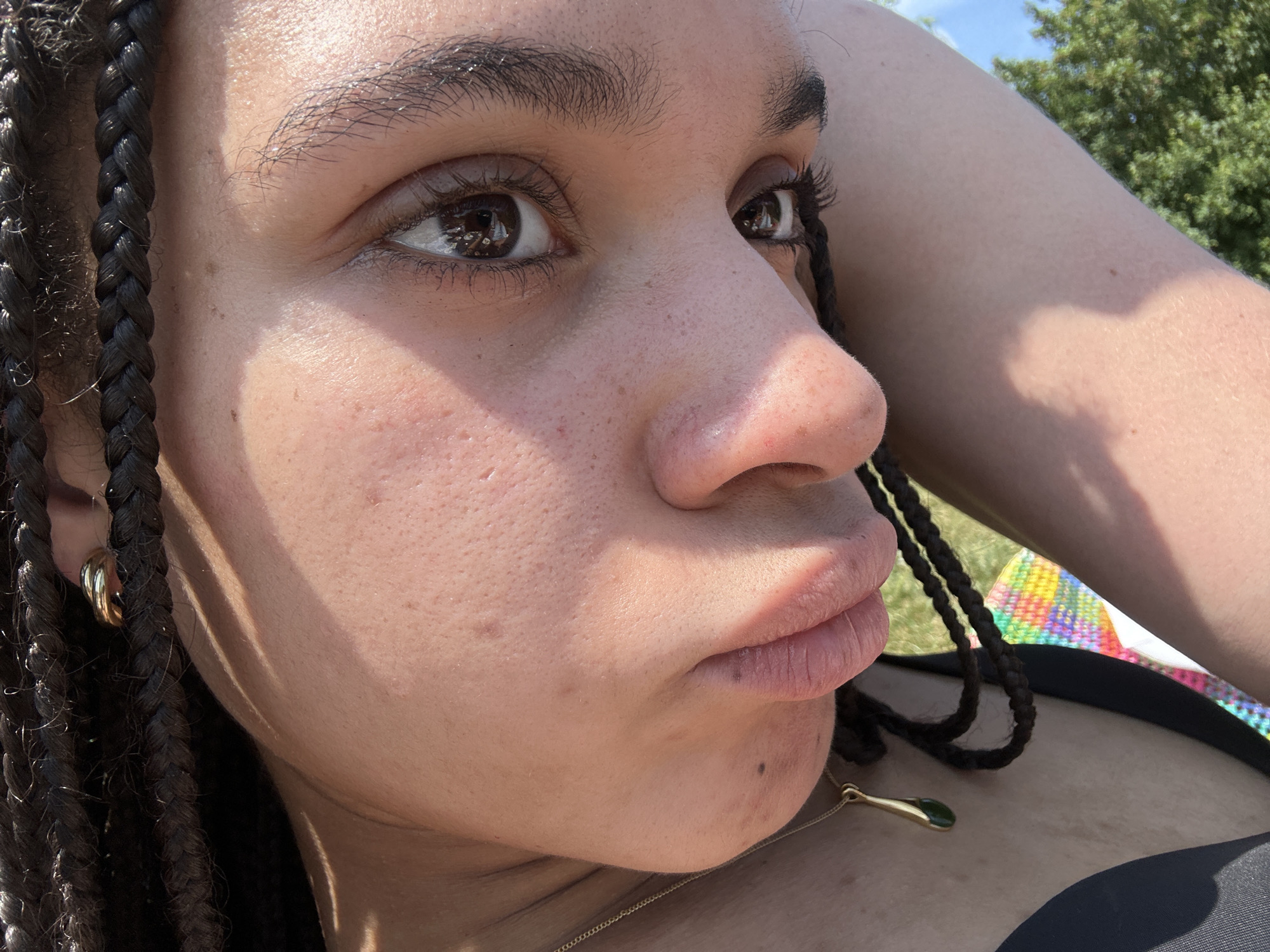 A close-up shot captures a young person with braided hair lying outside on a sunny day. The individual is positioned on their back, with the left arm raised behind their head. Their eyes gaze slightly upwards and to the right. The skin tone appears light brown, with visible pores and minor blemishes. A gold hoop earring is visible in the left ear. A gold necklace with a green pendant rests on the chest. A colorful blanket lies partially visible behind the subject's head, and green trees and a blue sky are in the background. The person appears to be lying on grass.