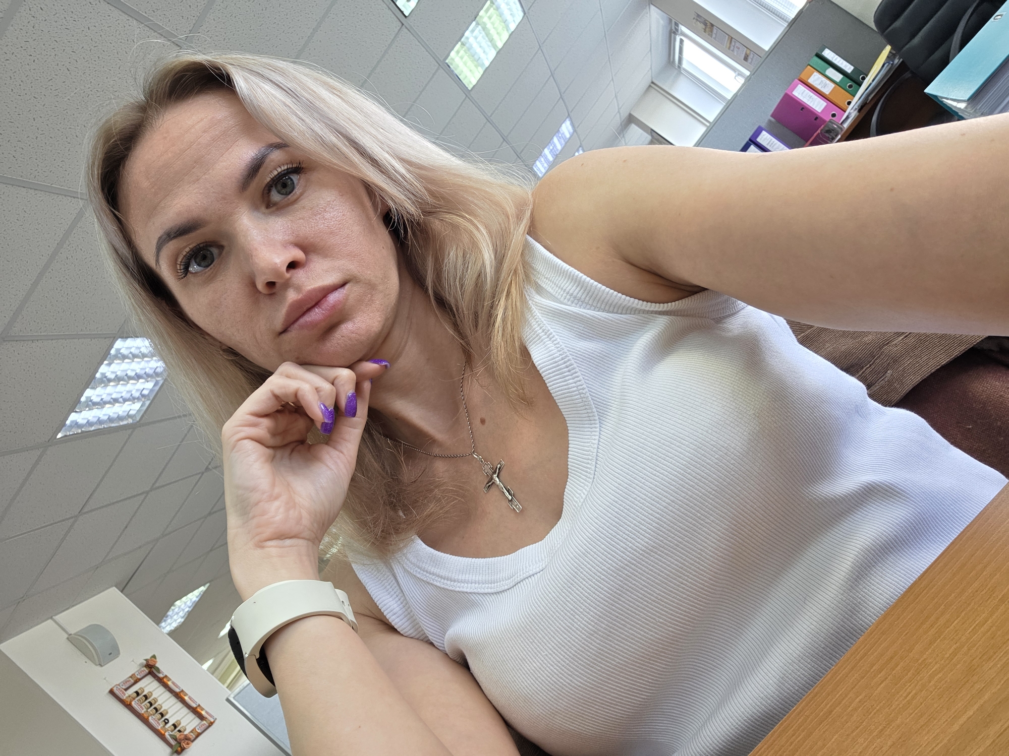 A fair-skinned woman with blond hair, gray eyes, and purple nail polish rests her chin on her hand. She is wearing a white tank top and a silver necklace with a cross. A white smartwatch is visible on her wrist. She is sitting at a wooden desk, and a brown chair is partially visible.
The location appears to be an office with ceiling lights, filing cabinets containing files of varying colors, and other office supplies. Based on the presence of the Russian abacus in the lower left corner, the location is likely Chaikovsky, Russia.