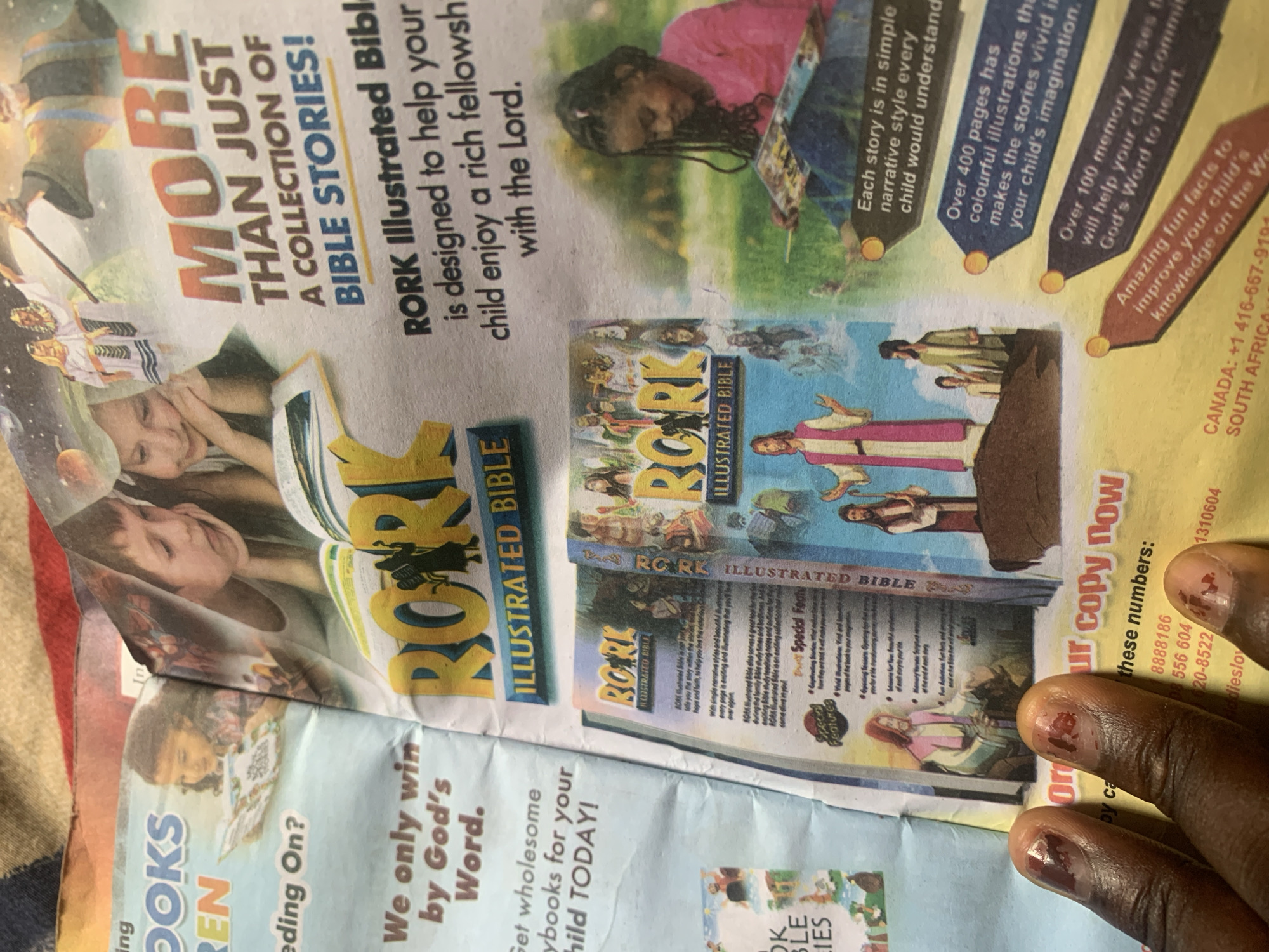 The image depicts a spread from a children's book advertisement, featuring vivid illustrations and promotional text. The dominant visual is a stylized title "RORK ILLUSTRATED BIBLE" in large, bold letters. Surrounding this are images of children reading, biblical scenes, and a young girl engrossed in a book. Text on the page highlights features like "More than just a collection of Bible stories!", "designed to help your child enjoy a rich fellowship with the Lord," and details about the number of pages and included memory verses. A call to action encourages readers to "Order your copy now" with phone numbers for Canada and South Africa. The bottom left corner shows a close-up of a person's hands with fingernails painted red. The image is a printed advertisement, not a scene from Jauro Yinu, Nigeria.