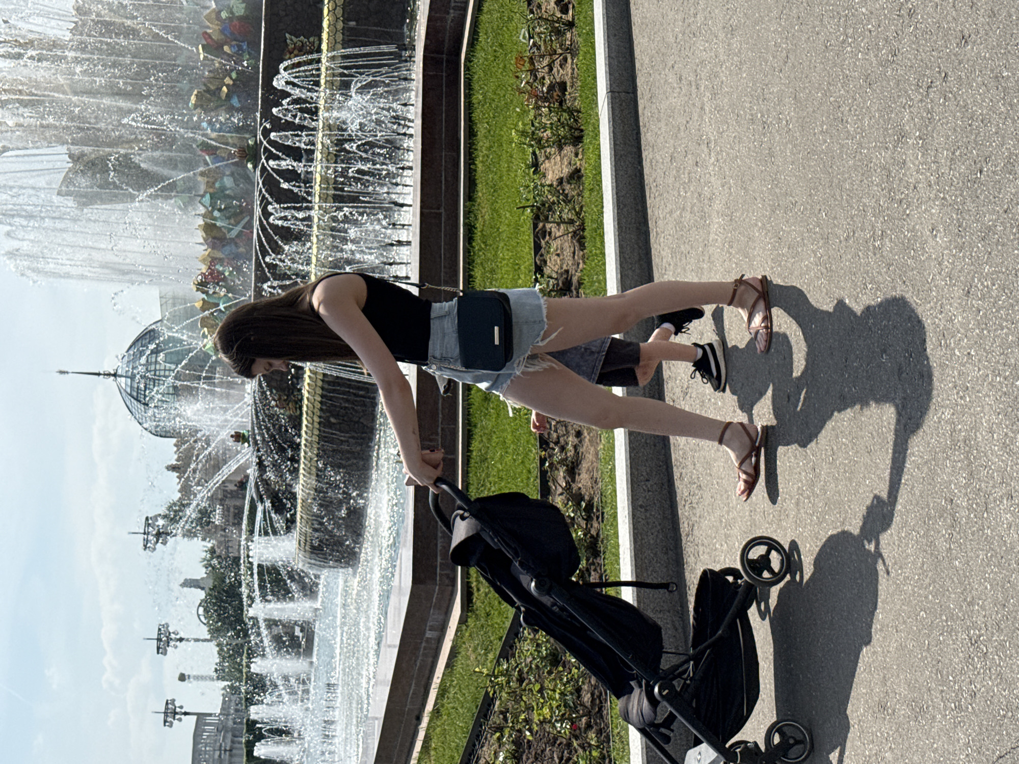 The image depicts a woman with long brown hair walking along a paved path next to a large fountain in Moscow, Russia. She is wearing a black tank top, denim shorts, and brown sandals. She is pushing a black stroller. Beside her, there is a small child wearing tennis shoes. The fountain is multi-tiered and elaborate, with numerous water jets. A green lawn and some bushes are present between the path and the fountain. The sky is partially visible. The image appears to be taken during the daytime.