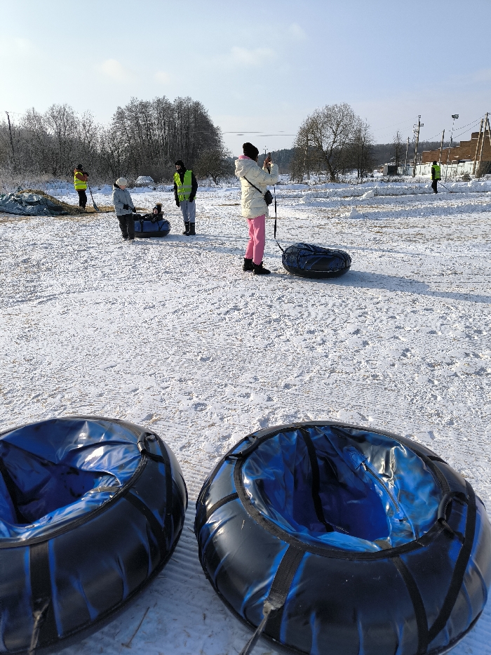 The image depicts an outdoor, snow-covered scene in Prybuzke, Ukraine, under a clear blue sky. In the immediate foreground, two black and blue inflatable snow tubes are visible. The midground features several individuals and objects: a person wearing a white jacket, pink pants, and a black hat stands, holding a smartphone in a horizontal orientation, appearing to take a photograph. An inflatable snow tube rests on the snow near this individual. Further back, a child is seated in a dark blue inflatable snow tube, attended by a person in a light-colored jacket. Nearby, another individual in a green high-visibility vest and dark pants stands. To the far left, a person in a yellow high-visibility vest is partially visible near a mound covered with a tarp, holding a shovel. To the far right, another person in a green high-visibility vest is also seen holding a shovel. The ground is covered in white snow with visible tracks and undisturbed areas. The background consists of a treeline with bare deciduous trees, utility poles, and distant structures, possibly residential or agricultural, under bright sunlight.