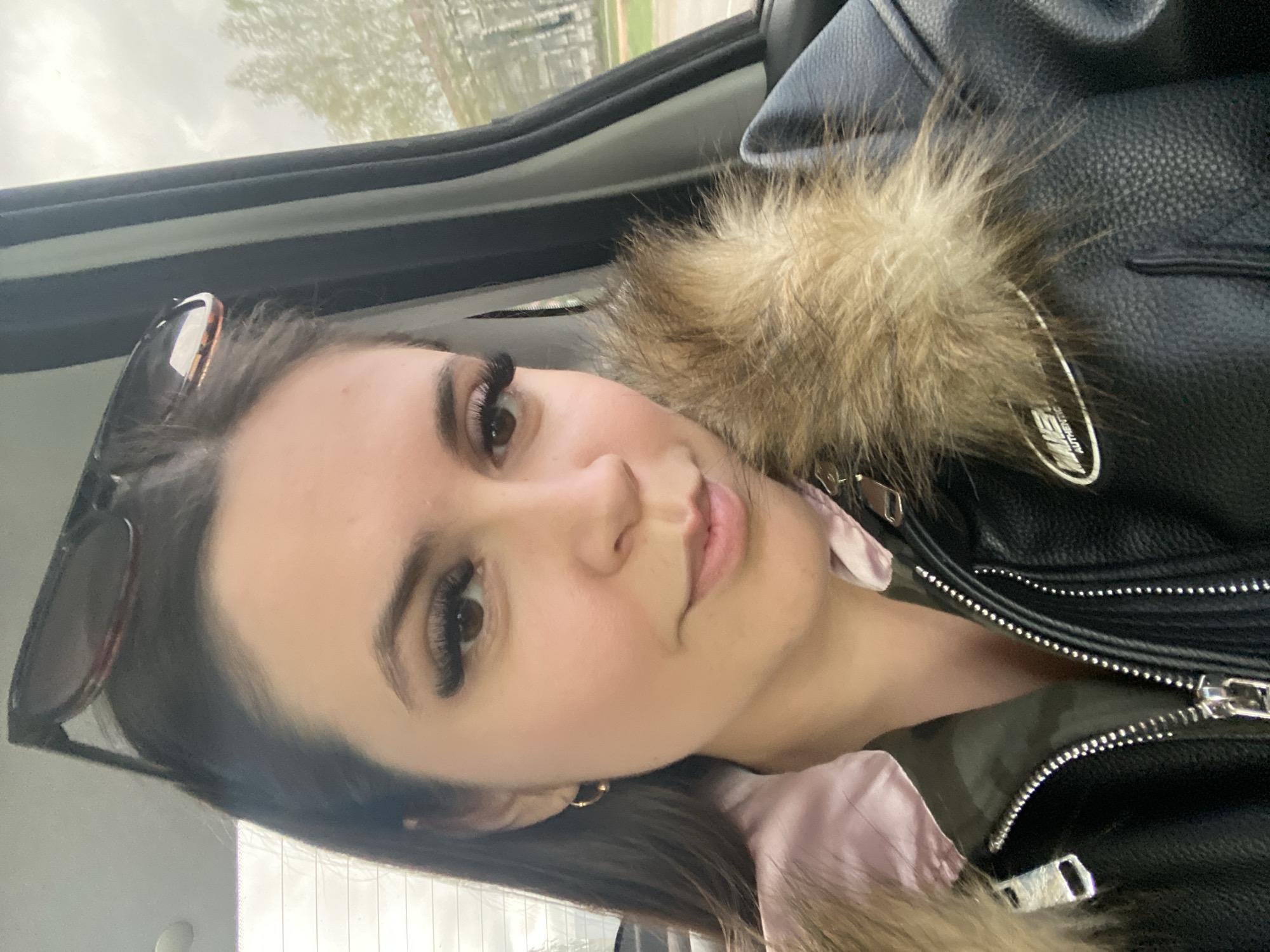 A woman is taking a selfie inside a car. She's wearing a black leather jacket with fur trim and sunglasses are perched on her head. Based on the surroundings visible through the car window, it is not possible to definitively confirm that the location is Chaikovsky, Russia. The environment outside the car window appears to be a typical urban or suburban landscape.
