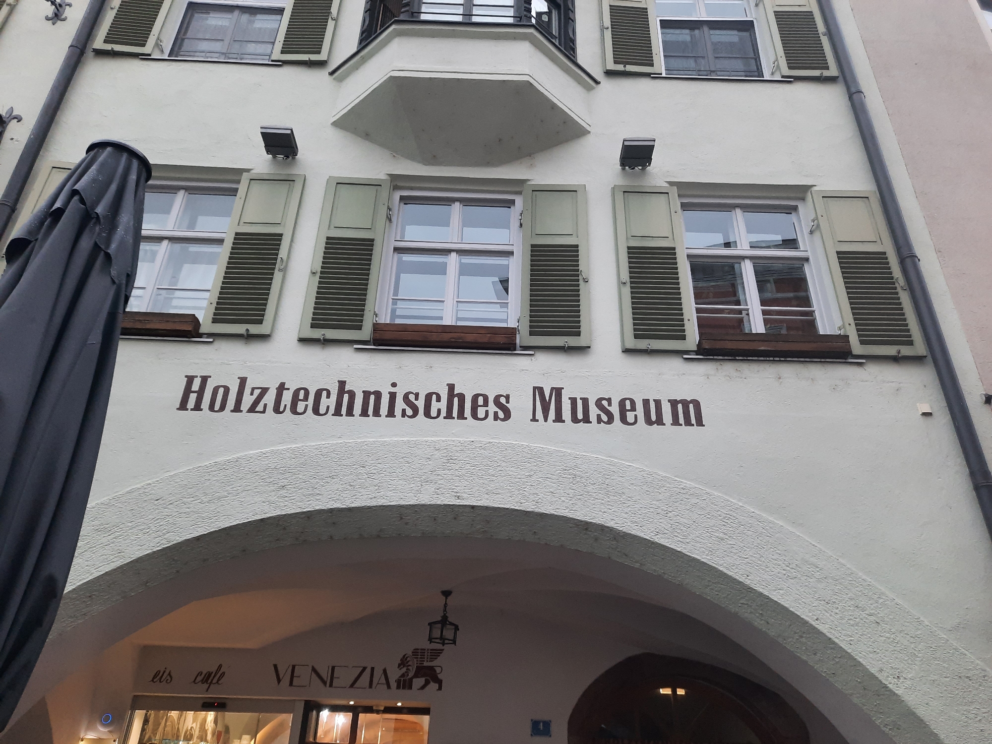 The image captures a building facade in Rosenheim, Germany, featuring the prominent signage "Holztechnisches Museum" above an arched entrance. The building is light-colored, with several windows adorned with green shutters. A dark, folded umbrella or awning occupies the left side of the frame, partially obscuring the building. Below the museum's name, the archway reveals the entrance to "eis cafe VENEZIA," identifiable by its signage and a lion emblem. The scene appears to be an exterior view of a commercial and cultural establishment within the city.