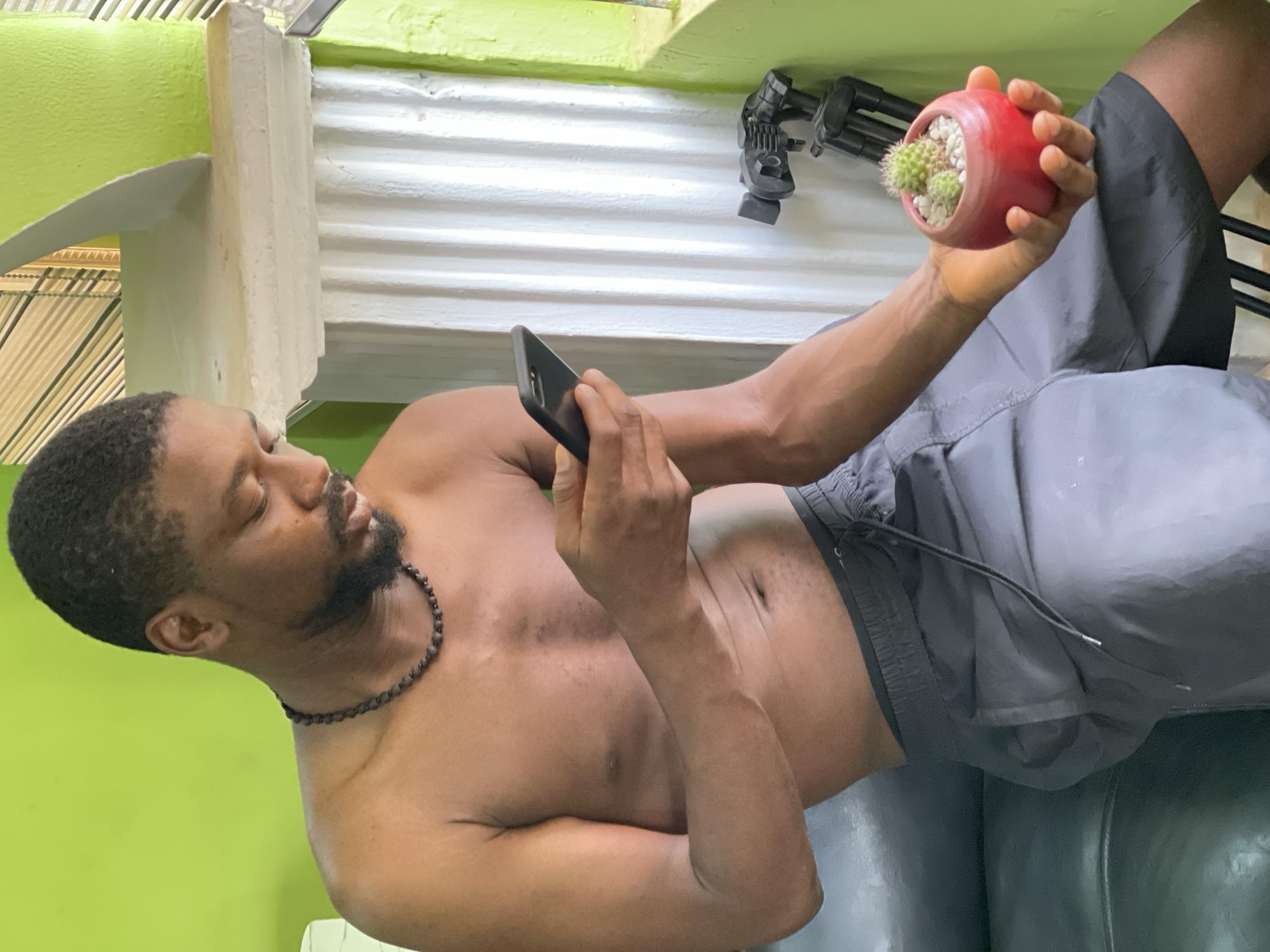 A shirtless man with dark skin, a beard, and a beaded necklace is reclined on a dark, possibly leather, piece of furniture. He's holding a black smartphone in one hand and a small, red pot containing a cactus in the other. The background features a bright green wall, a white corrugated surface, and a camera tripod. The scene takes place indoors. It is impossible to determine if the location is in Abuja, Nigeria.