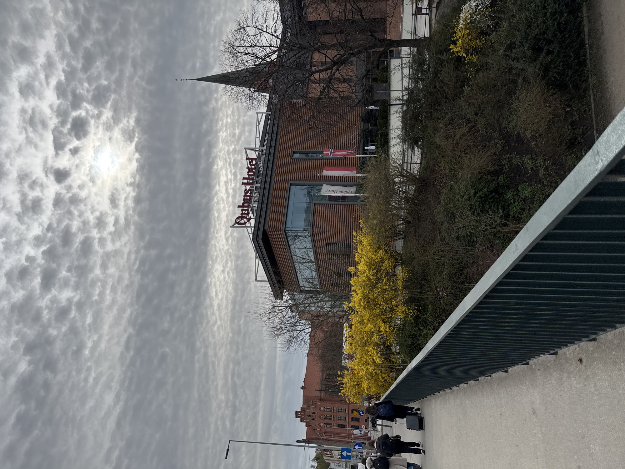 The image shows a view in Bydgoszcz, Poland, from a bridge, likely over the Brda River. On the right is the bank of the river with the "Qubus Hotel" building featuring a brown wooden facade. Two flags, including the Polish flag, are visible in front of the hotel. To the left of the hotel, a church steeple rises above the building. In the foreground, people are walking along the bridge, some pulling luggage. The sky is overcast with a thin cloud cover. A metal railing lines the right side of the bridge. A yellow flowering bush grows along the riverbank. In the distance, brick buildings can be seen. A street lamp and traffic signs are also visible in the lower left corner.