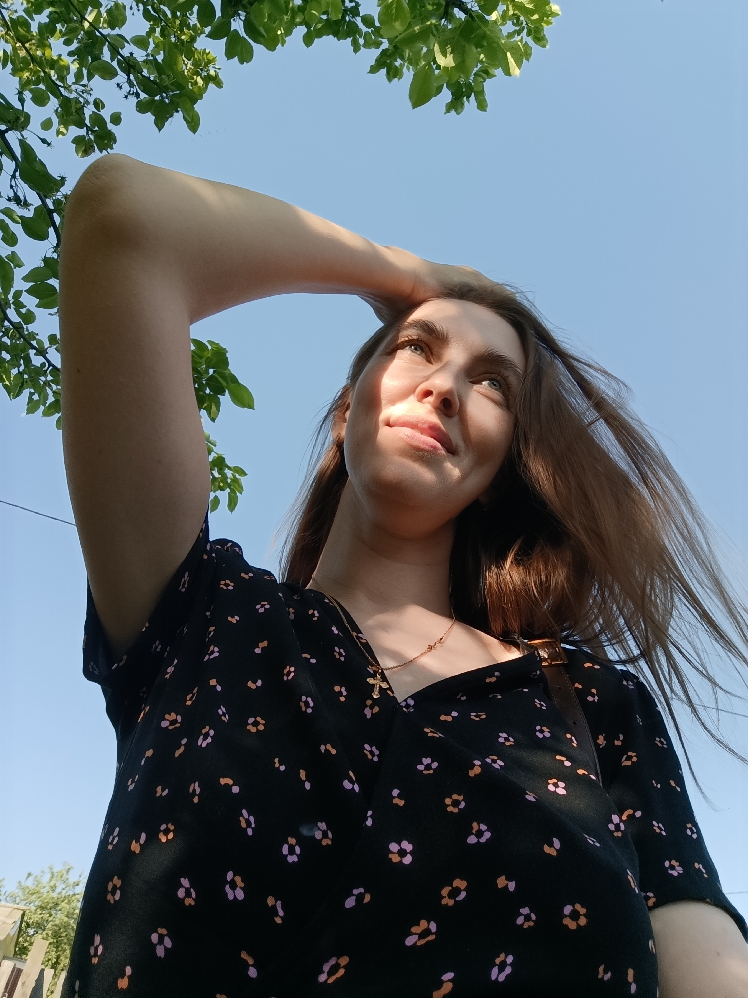 A low-angle shot captures a woman outdoors. She is fair-skinned with long brown hair, and is wearing a black floral-patterned dress and a gold necklace. Her left arm is raised, with her hand touching her head. She looks upwards with a slight smile. The background consists of a clear, bright blue sky, with a small section of green leaves visible in the top left corner. A fence and more foliage are visible along the lower edge of the image. The location is the city of Chaikovsky, Russia.