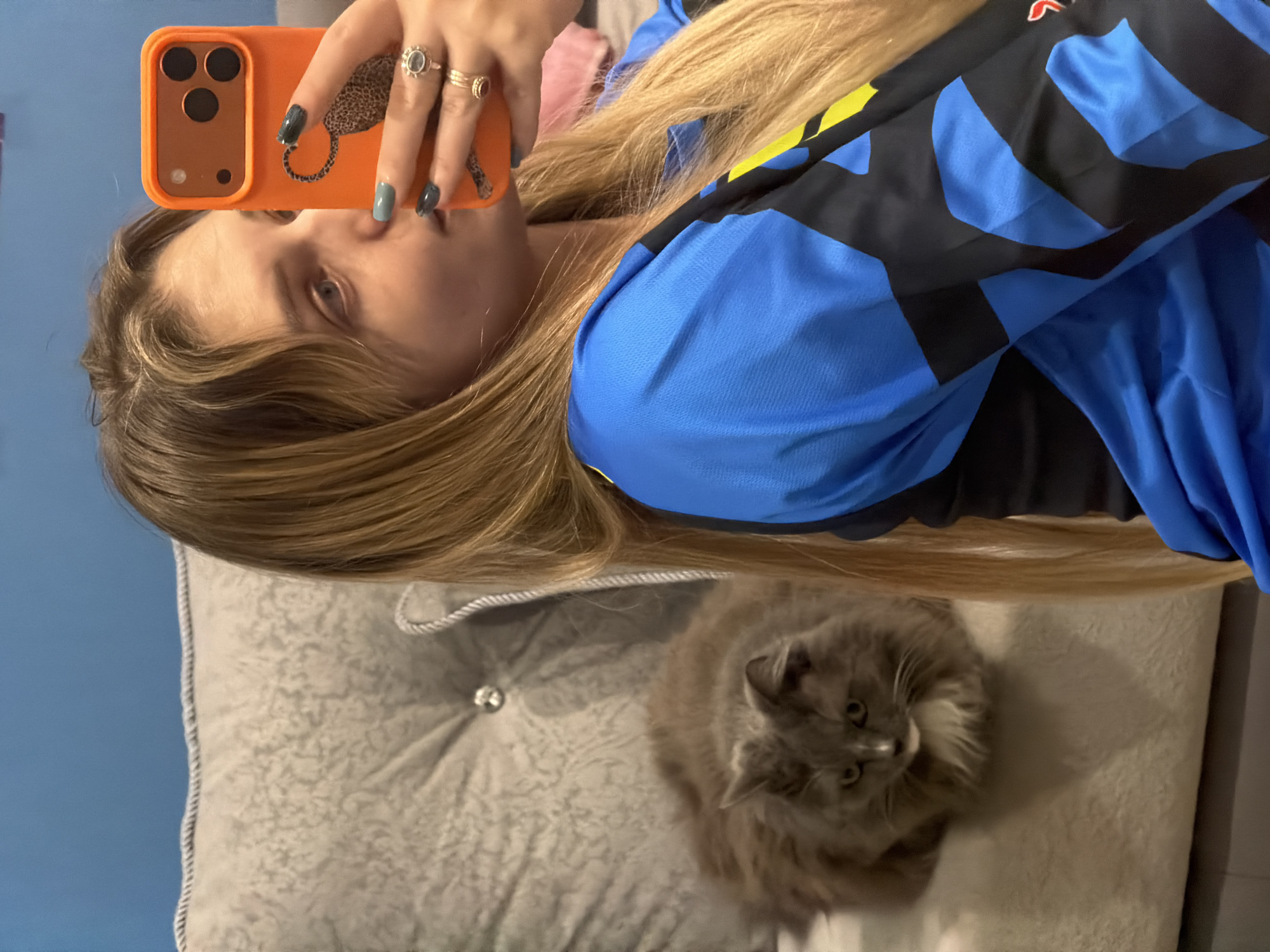 A young woman is seen from a slightly elevated, angled perspective, holding an orange smartphone up to take a selfie. The phone has a leopard print design on the back. She is wearing rings on her fingers and has her nails painted in different shades of blue and gray. She has light brown hair and is wearing a blue and black top with yellow accents. She is lying on a gray, textured pillow with a button. Next to her on the pillow is a gray, long-haired cat looking up towards the camera. The background is a plain blue wall. This photograph does not permit identification of any specific location in the city of Большая Шильна, Russia.
