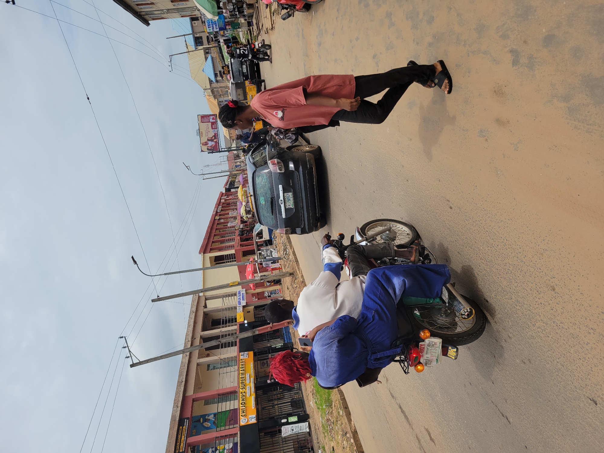 In Egbagu, Nigeria, on a dusty road, two people ride a motorcycle. The passenger, a woman with bright red hair and a blue dress, holds a smartphone. The driver is a man in a white shirt. A black Toyota Matrix is parked on the side of the road. A young woman in a pink t-shirt and black pants walks away from the camera. Buildings with shopfronts line the street, featuring signs for a supermarket and a pharmacy. Overhead, power lines stretch across the sky, which is partly cloudy. The overall scene depicts a typical street view in a bustling Nigerian city.