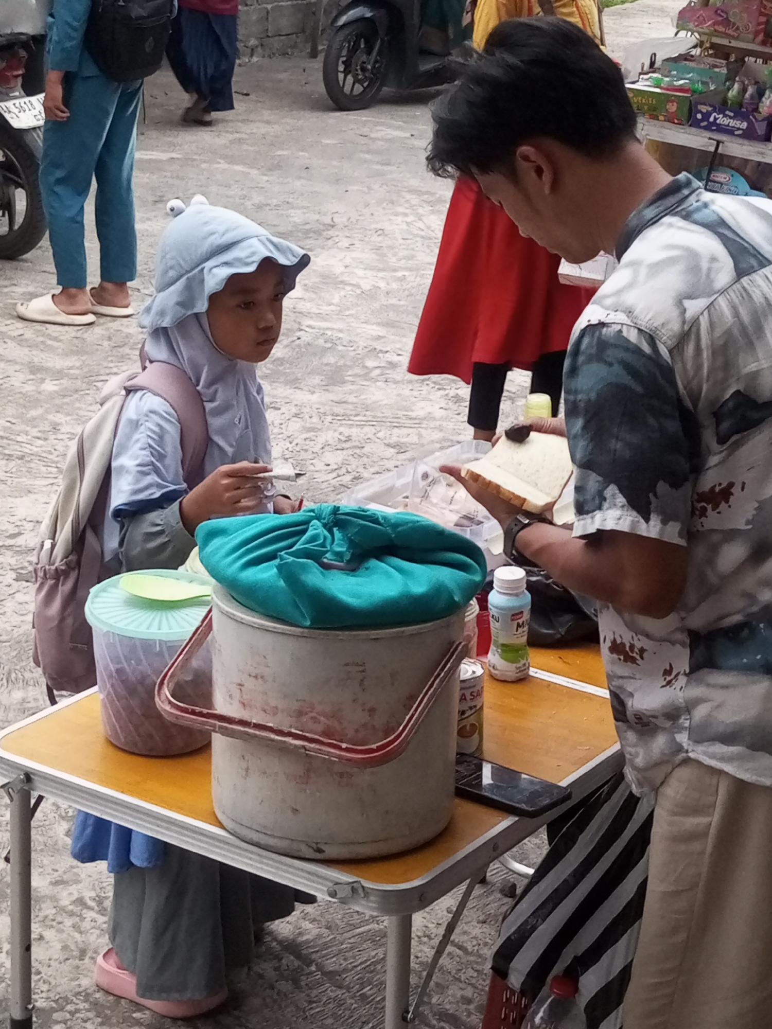 A young person wearing a blue hijab and a light blue outfit with a backpack is seated at a yellow folding table outdoors, holding a piece of white bread. They are looking towards an adult male, who is standing and holding another slice of bread with chocolate spread on it. The adult is wearing a patterned shirt and beige pants. On the table are two buckets, one covered with a turquoise cloth and another a lighter color, a plastic container with a green lid and spoon, and a bottle of white liquid with green labeling. In the background, a motorcycle is partially visible, along with other people walking and standing. The ground is a rough, light-colored surface, possibly concrete or asphalt. A sign with Indonesian text and numbers is attached to the motorcycle. The scene appears to be in a market or street area, potentially in Krasak, Indonesia, given the context provided.