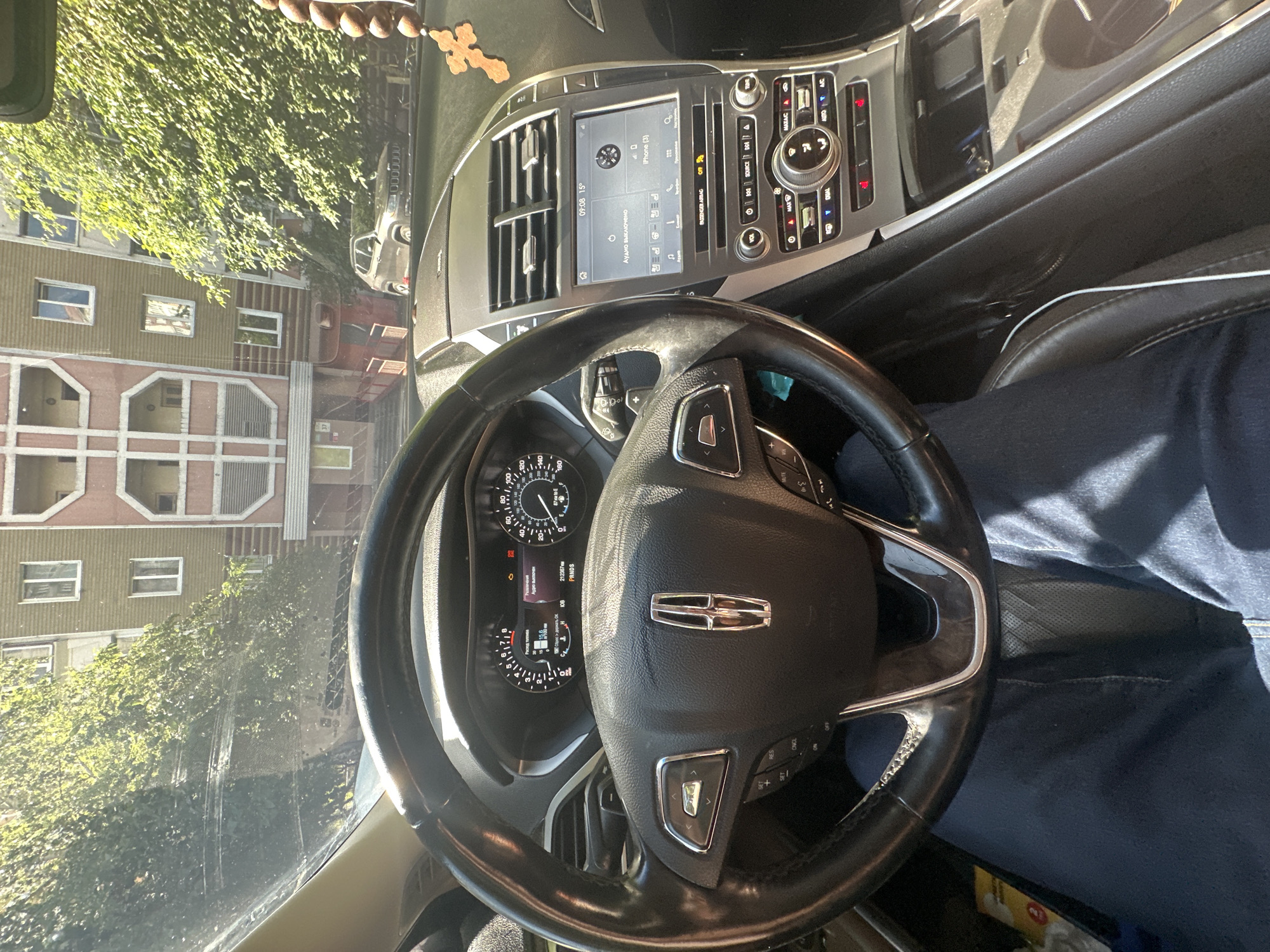 The image shows the interior of a car, likely a Lincoln, parked on a sunny day in front of an apartment building. The steering wheel, dashboard, and infotainment screen are visible. The infotainment screen displays the time as 09:08 and a temperature of 15 degrees. It also indicates that the iPhone is connected and shows a Bluetooth symbol. Outside the car, through the windshield, a multi-story apartment building with beige and pink facades is visible, along with leafy green trees providing shade. A silver SUV is parked on the street in front of the building. The car appears to be parked on a street in Kyiv, Ukraine, given the architecture and vegetation. A wooden cross pendant hangs from the rearview mirror. The driver's legs, clad in blue jeans, are visible in the foreground.