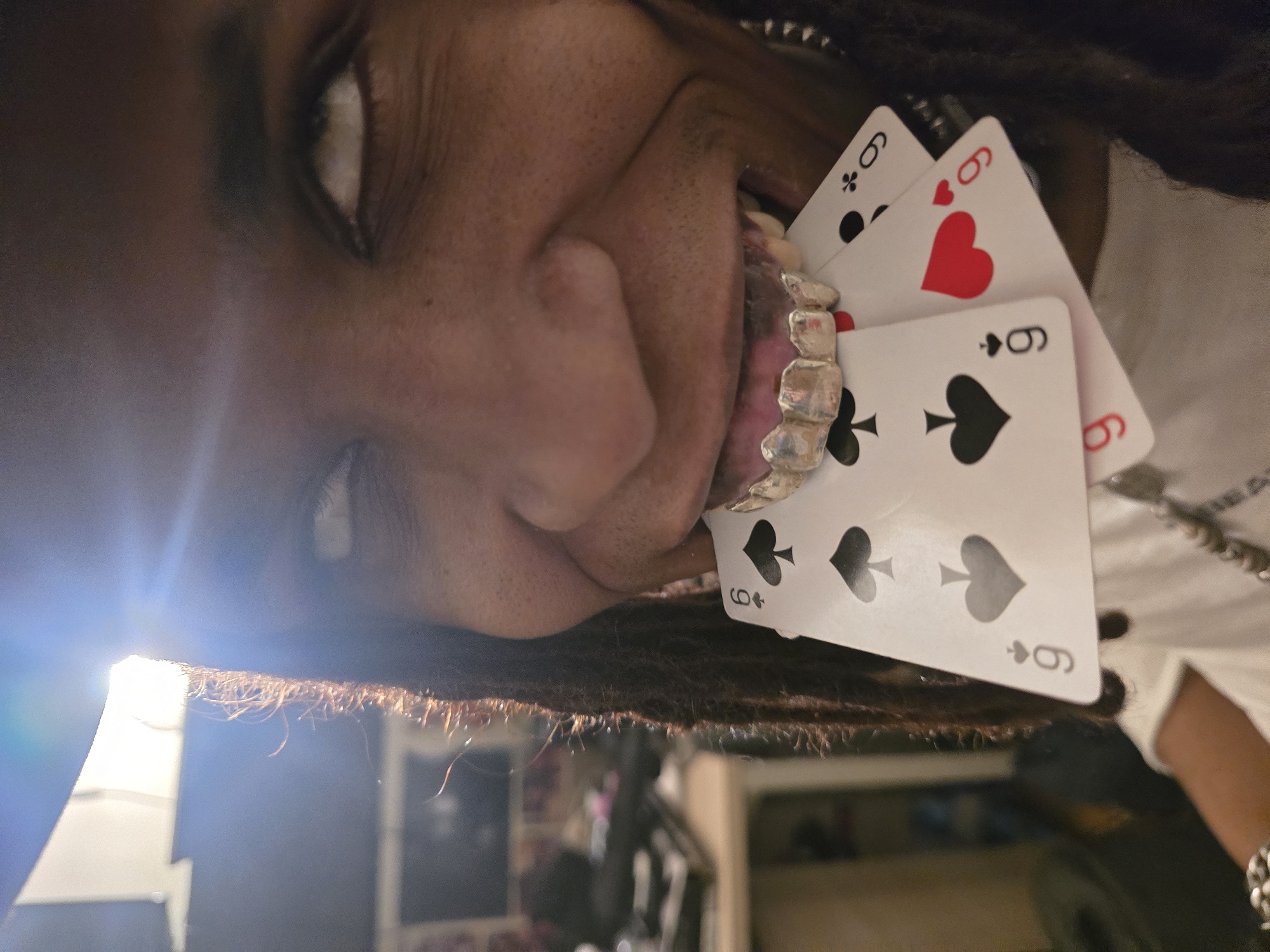 The image shows a person with dark skin, dreadlocks, and silver grillz on their teeth holding three playing cards in their mouth. The cards are all nines, with different suits: clubs, hearts, and spades. The person's face is close to the camera, and their eyes are visible. The background is blurry but appears to be an interior space. There is no indication of a location in Paris, France.