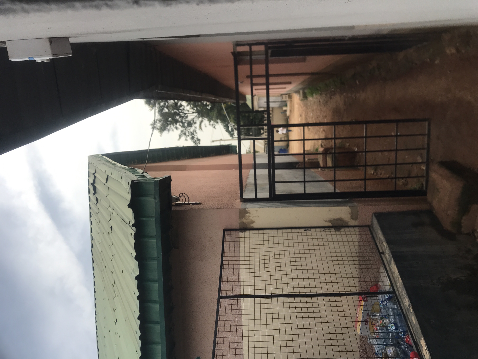 The image depicts an outdoor scene, possibly in Jos, Nigeria, with two buildings that appear to be adjacent, creating a narrow passageway between them. The building on the left has a pinkish-red textured wall and a green corrugated metal roof. A black metal gate with a grid pattern is visible, and behind it, a large wire mesh enclosure contains a pile of plastic bottles and what appears to be a cardboard box. The building on the right also has a pinkish-red wall and a dark, sloped roof or overhang. The ground in the passageway is dirt. In the distance, a person in a white shirt is walking away from the camera. The sky is overcast with clouds. A white electrical box is mounted on the exterior wall of the building on the right, near the top of the image.