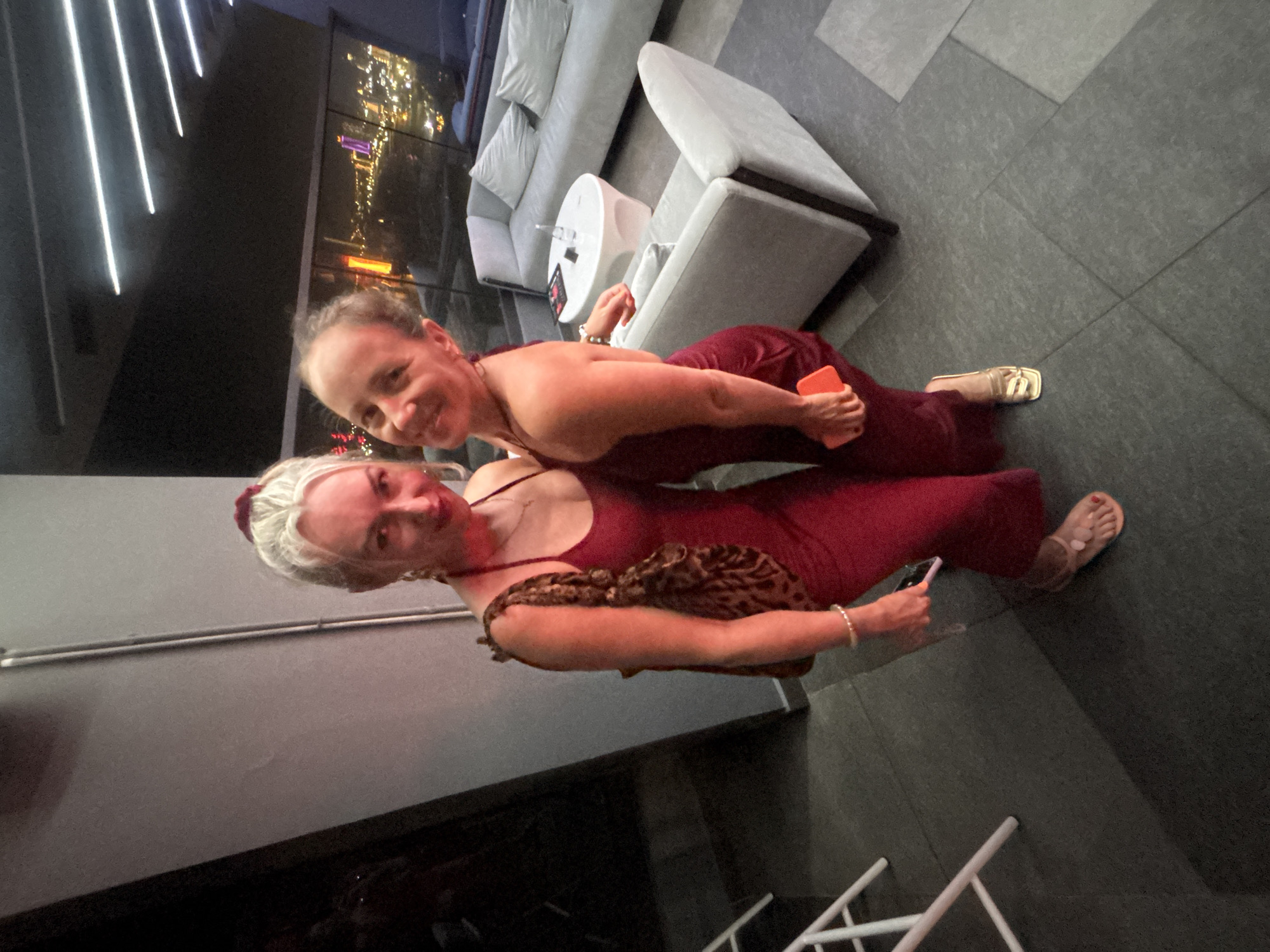 Two women are posing for a photo indoors. The woman on the left has blonde hair styled in a bun with a red scrunchie and is wearing a red dress, a leopard print purse, and a pearl bracelet. The woman on the right has brown hair and is wearing a red dress and gold sandals. She is holding an orange phone. They are standing on a dark gray tiled floor. In the background, there is a seating area with a white table and light gray sofas, and a large window overlooking a city at night, which is likely Đà Nẵng, Vietnam, given the vibrant lights and waterfront structures visible. The overall lighting is dim, with artificial lights from the ceiling and the city outside illuminating the scene.