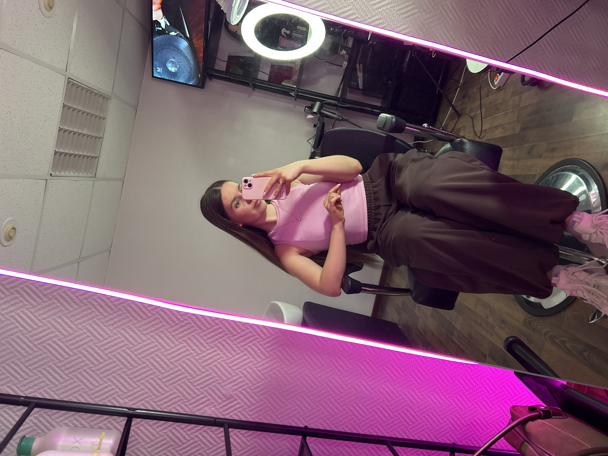 A young woman is posing for a mirror selfie in what appears to be a beauty salon or studio. She is sitting in a stylist's chair, wearing a pink top and brown trousers, and holding a pink smartphone. The mirror is outlined with bright pink LED lighting. The background shows salon equipment, a TV screen, and product shelving. The location is Klin, Russia.