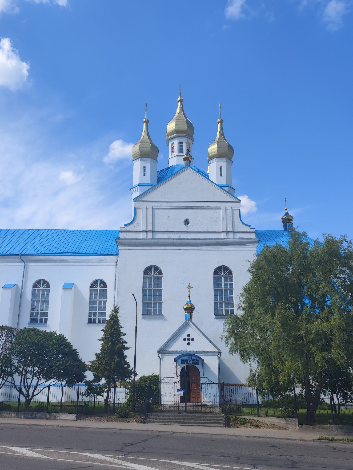 The image shows a white church with a blue roof and golden domes, located in the city of Slonim, Belarus. The church has a symmetrical facade with arched windows and a central entrance. The entrance is adorned with a small golden dome and a cross. Trees and a black metal fence surround the church. The scene is captured from a street-level perspective, with a road and crosswalk visible in the foreground. The sky is blue with scattered clouds, suggesting daytime.