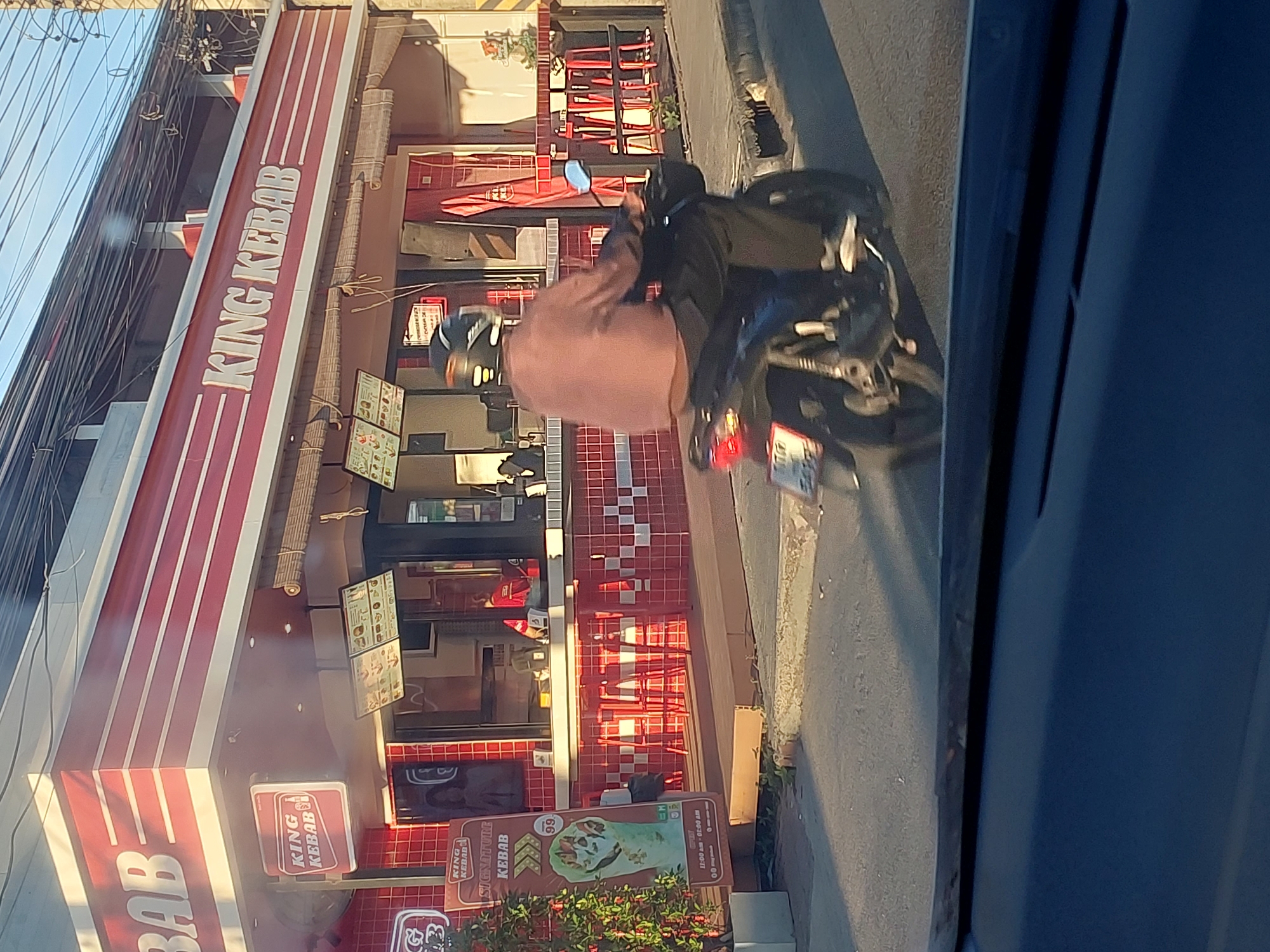This media file captures a street view of a "King Kebab" restaurant in Baan Bo Phut, Thailand. The scene is dominated by the red and white striped facade of the restaurant, with its prominent signage. In the foreground, a person wearing a light pink shirt and a helmet is riding a motorcycle, with another person partially visible behind them also wearing a helmet. The motorcycle's brake light is illuminated. The time of day appears to be late afternoon or early evening, suggested by the warm, angled sunlight casting shadows.
Various signs and displays are visible around the restaurant, including menus featuring food items and prices. The restaurant itself appears to be open, with seating visible through the windows. The street outside is paved, and there are some plants growing near the establishment. Electrical wires are strung above the scene, a common sight in many parts of Thailand. The overall impression is one of a busy urban street with a focus on street food. Visible text includes "KING KEBAB", "SIGNATURE KEBAB", and hours of operation "11:00 am - 01:00 am".