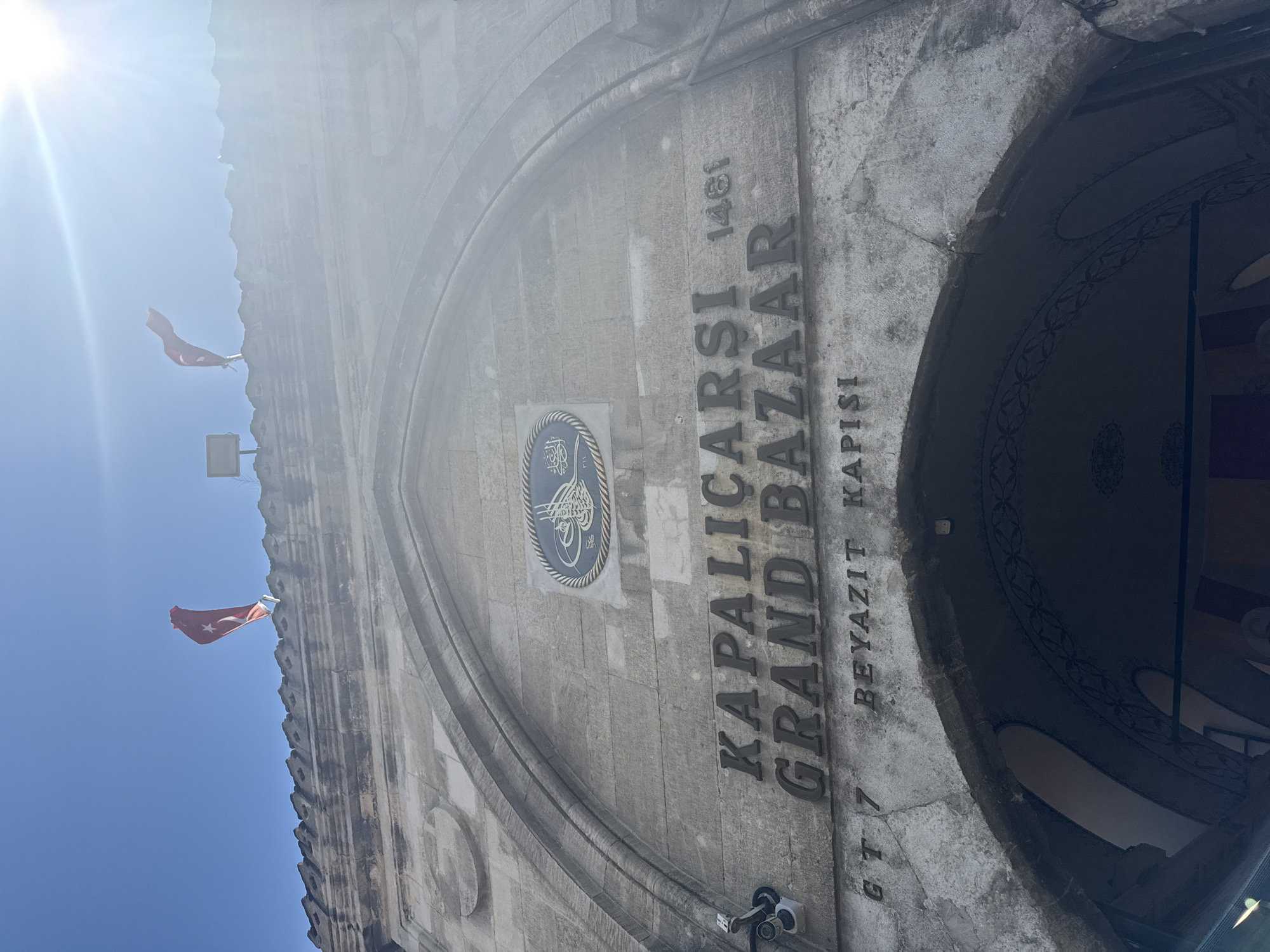 The image depicts the Beyazit Gate entrance of the Kapalıçarşı (Grand Bazaar) in Istanbul, Turkey. The primary subject is a large, weathered stone archway forming the entrance to a historical structure. Prominently displayed on the facade are the carved words "KAPALIÇARŞI GRAND BAZAAR" with the year "1461" adjacent to "ÇARŞI". Below, the text "BEYAZIT KAPISI" is visible, along with "G T 7" to its left. An oval plaque featuring intricate calligraphic script is mounted above the main text, centered within the arch's curve.
On the roofline above the archway, two red Turkish flags, each featuring a white crescent moon and star, are visible against a clear blue sky, appearing to wave in a breeze. A security camera is affixed to the stone wall on the left side of the archway, pointing downwards. A floodlight or similar illumination fixture is also visible near the left flag on the rooftop. Intense sunlight from the upper right creates significant lens flare, particularly affecting the upper right quadrant of the image and partially obscuring details on that side of the structure. The interior of the archway is dimly lit, revealing faint decorative patterns on its ceiling. No individuals are discernible within the frame.