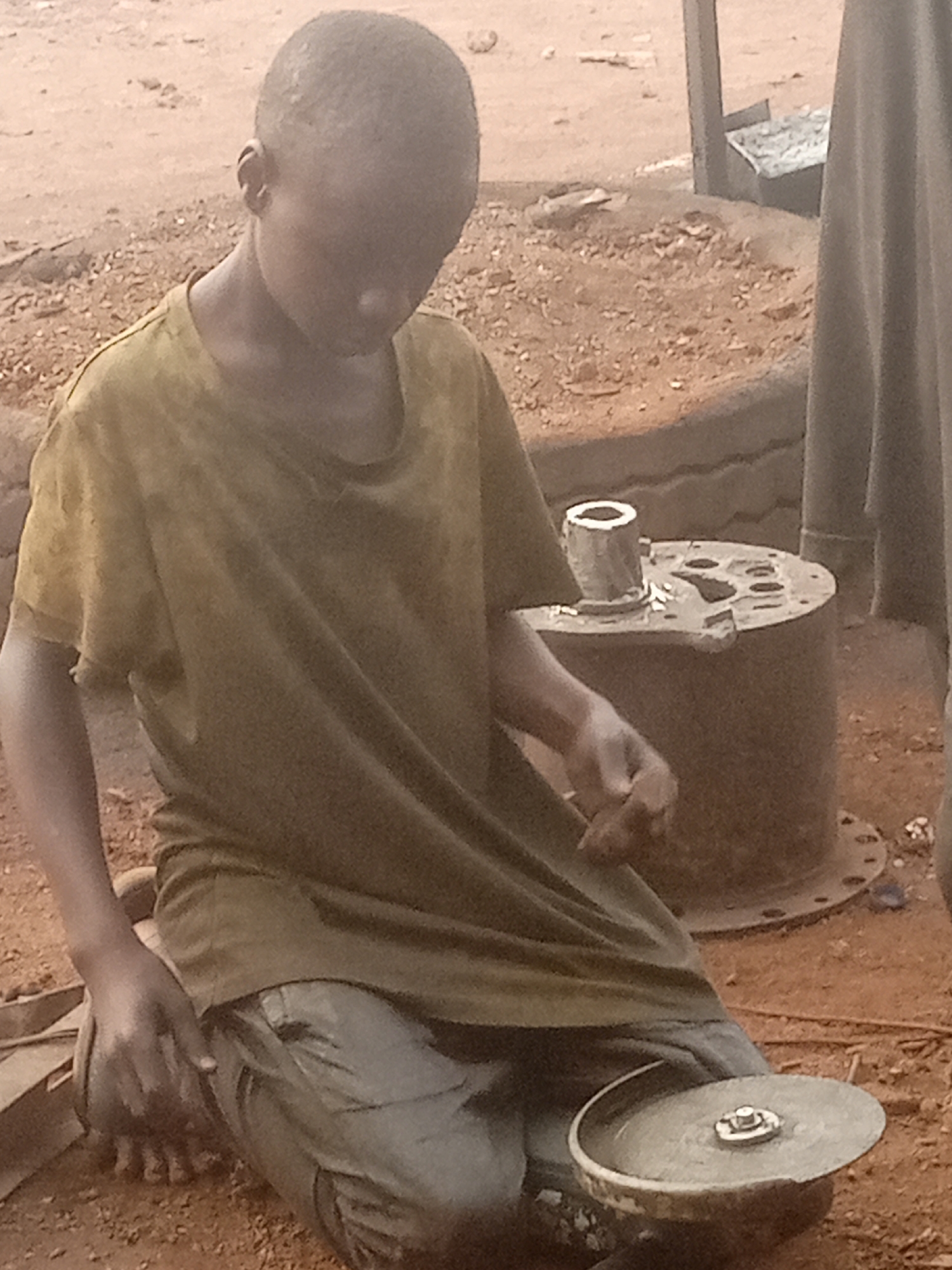 A child kneels outdoors in Bukuru, Nigeria, grinding an object with a grinding wheel. The child, wearing a brown t-shirt and grey trousers, is surrounded by red dirt. Next to the child is a metal cylinder with holes. In the background, there is what looks like the edge of a tire and some metal pipes.