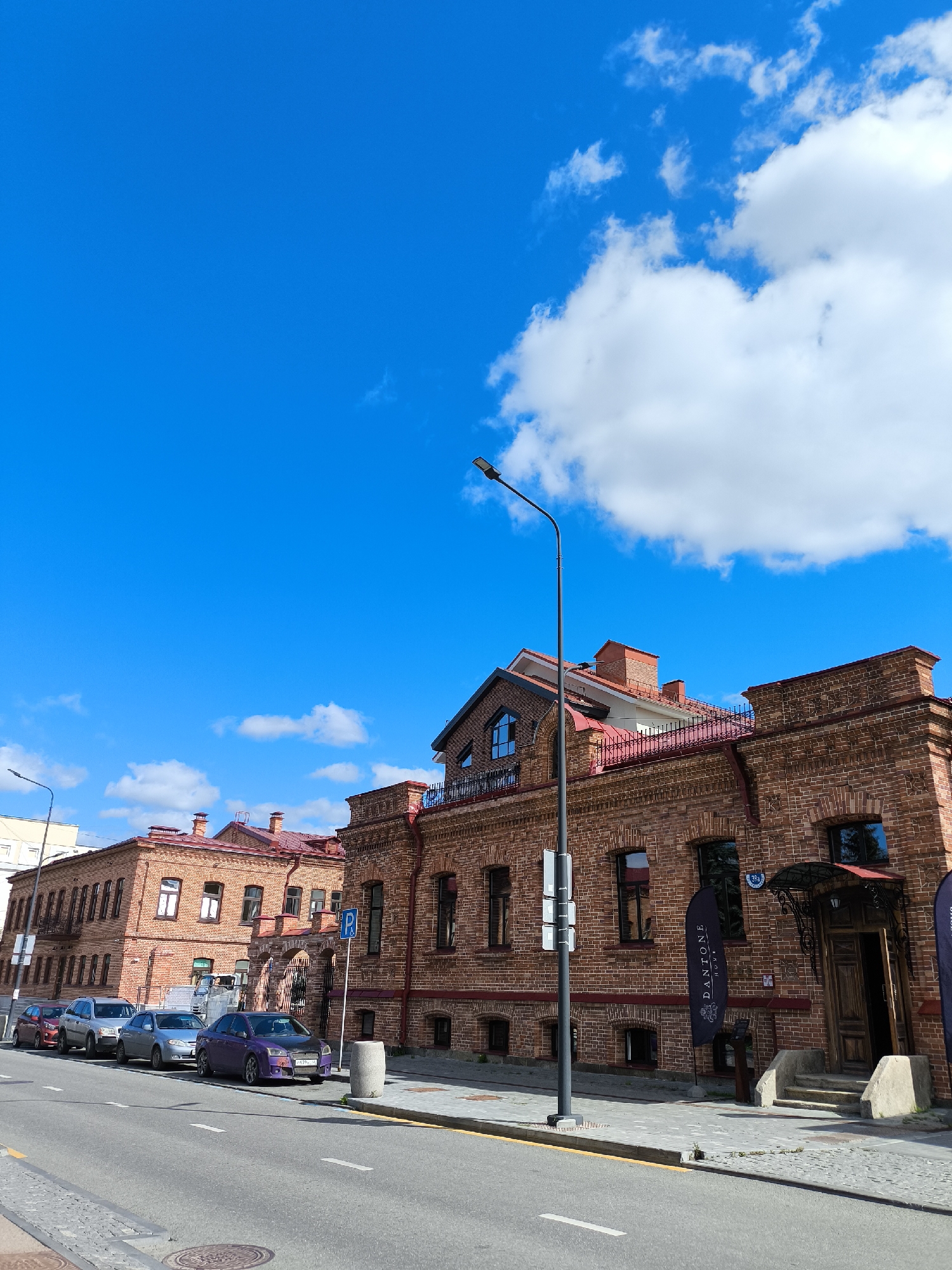 The image shows a street scene in Yekaterinburg, Russia, featuring red brick buildings and parked cars under a bright blue sky with scattered clouds. The buildings appear to be historic, with arched doorways and multiple windows. One building has a sign that reads "DANTONE". There are several cars parked along the street, including a purple car in the foreground. A lamppost stands on the sidewalk, and a trash can is visible near the building entrance. The street itself is paved with asphalt and bordered by a sidewalk made of cobblestones.