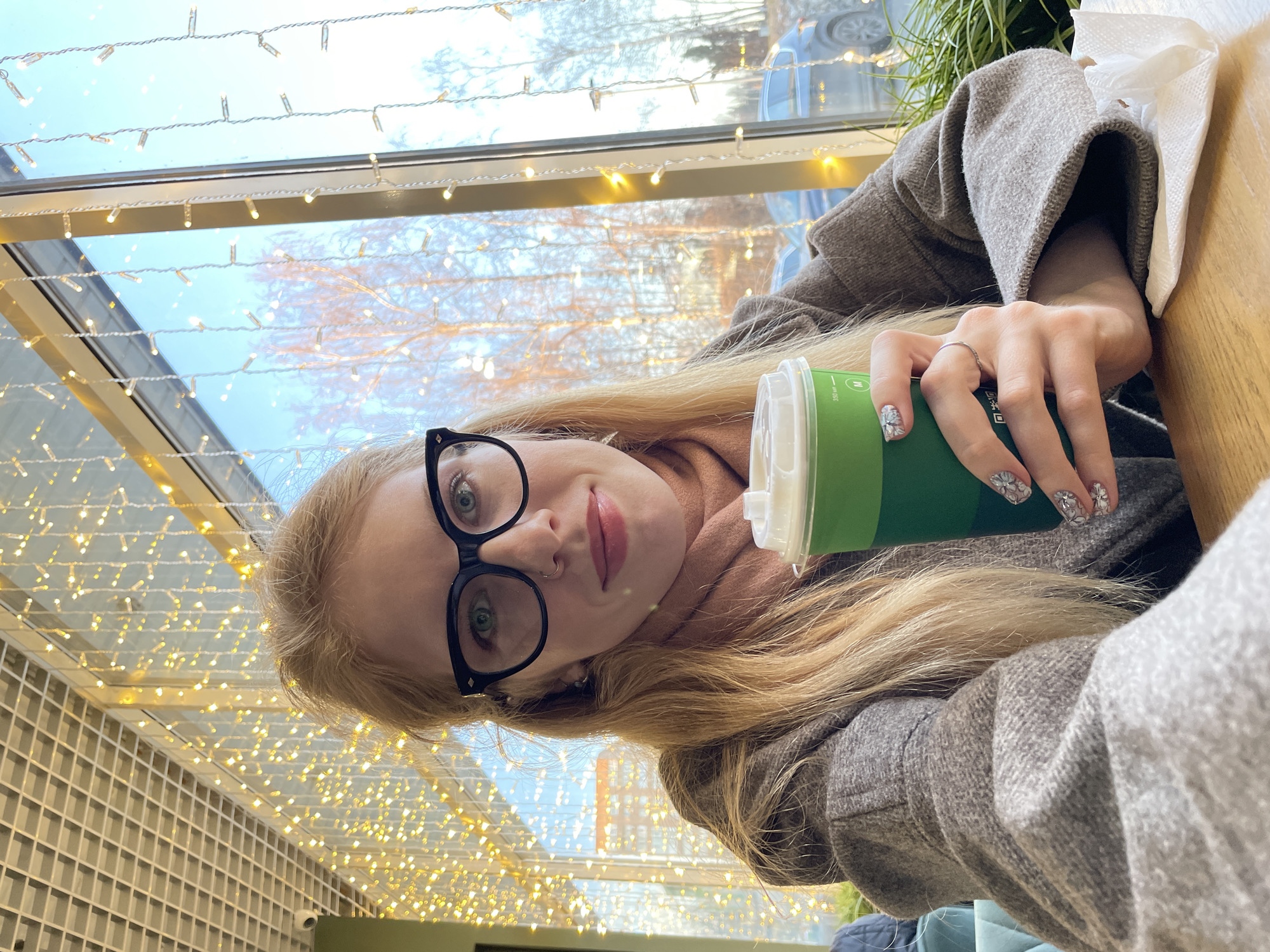 A woman with fair hair and glasses is holding a green paper cup in what appears to be a cafe. The cafe is decorated with fairy lights. She is wearing a grey coat or jacket. The location is likely Izhevsk, Russia based on details in the window's reflection. A light-colored building is visible.