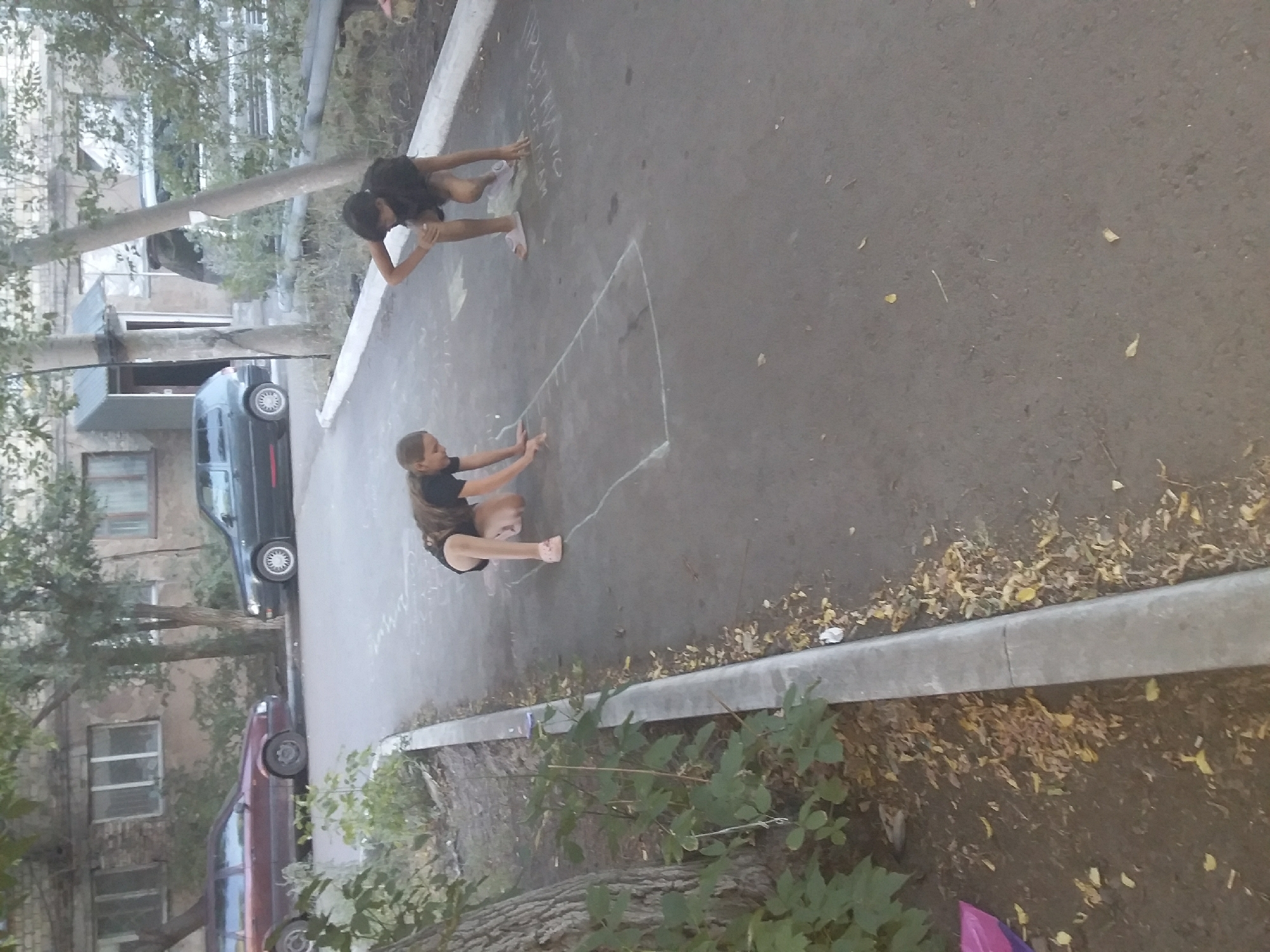 Two young girls are playing hopscotch on a paved surface in an urban environment. The girl on the left is on her hands and knees, positioned within one of the chalk-drawn squares. The girl on the right is in a crouched position, with one knee on the ground and the other foot flat, her hands extended forward. Both girls are wearing casual clothing. The hopscotch grid is drawn with chalk on the asphalt. In the background, there is a building with windows and trees. There are also parked cars visible, a dark grey hatchback and a maroon station wagon. The ground is an asphalt pathway with a concrete curb and some scattered leaves. The overall scene suggests a warm day, possibly late afternoon, given the lighting. The location is identified as the city of Saran, Kazakhstan.