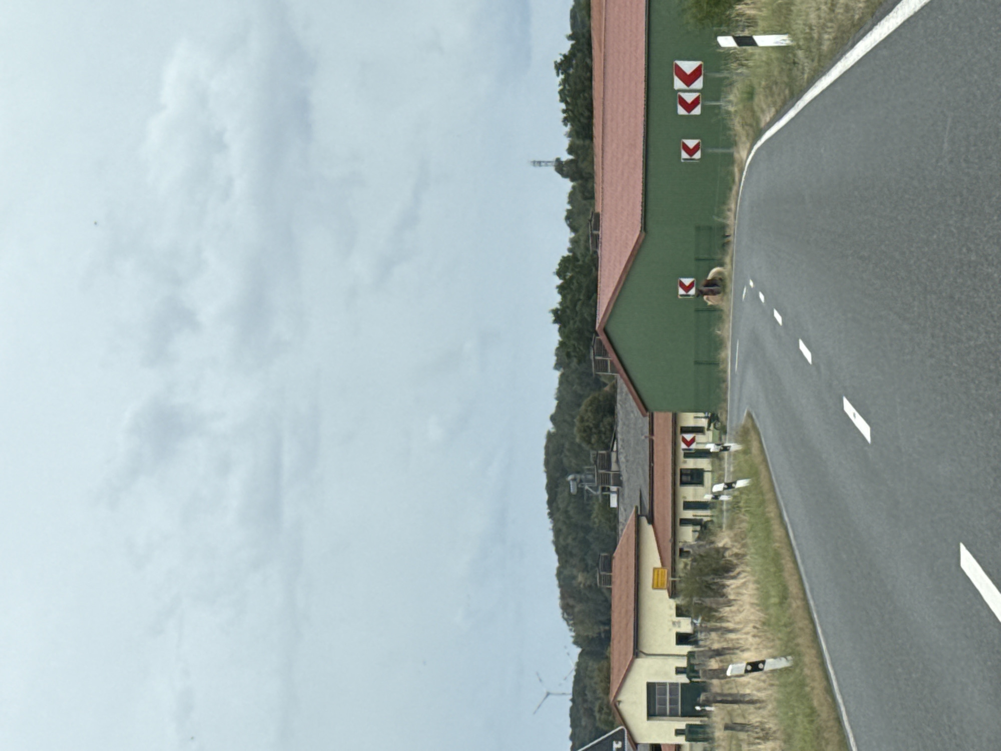 The image shows a rural road curving to the right in Hohenstein, Germany. On the left side of the road, there are several buildings with red roofs, including a yellow building with a garage door. A fence runs along the edge of the property. On the right side of the road, there is a large green building with a red roof. Several traffic signs indicating curves are visible along the road. In the background, there are rolling hills covered in trees. The sky is overcast. There are no people visible in the image.