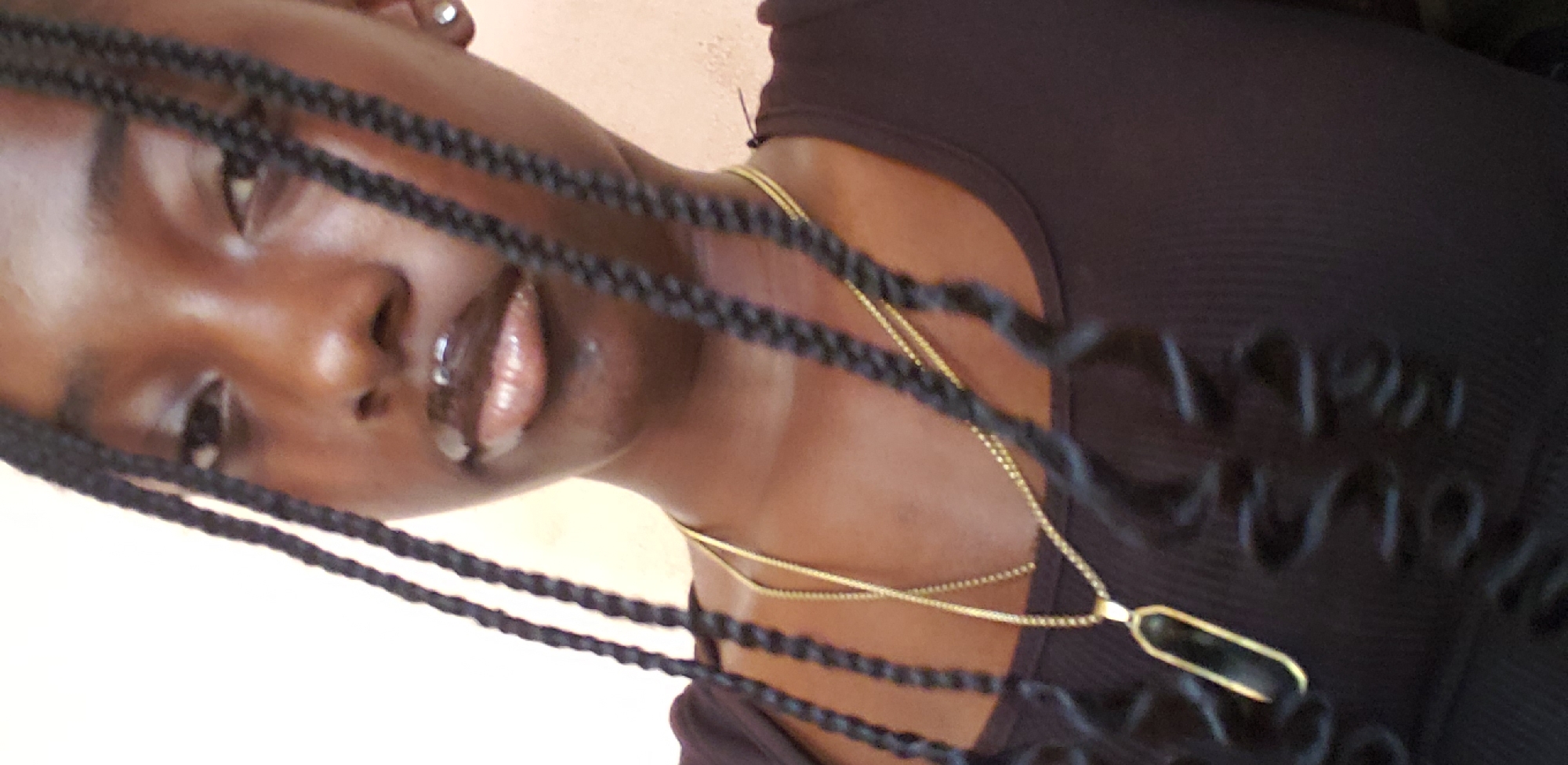 A close-up, tilted portrait features a young person with dark skin, looking upwards and slightly to the right with a soft, open-mouthed expression and glossy lips. Their face is framed by numerous thin, dark braids. They wear a dark, possibly ribbed, top and two layered gold necklaces. The upper necklace has an elongated, dark, possibly stone, pendant set in gold. A small silver stud earring is visible in the left ear. The background is a plain, light-colored surface, suggesting an indoor setting. The natural lighting indicates it is daytime. The subject appears calm, possibly posing for a selfie. There is no visible text in the image. The location context is Rido, Nigeria.