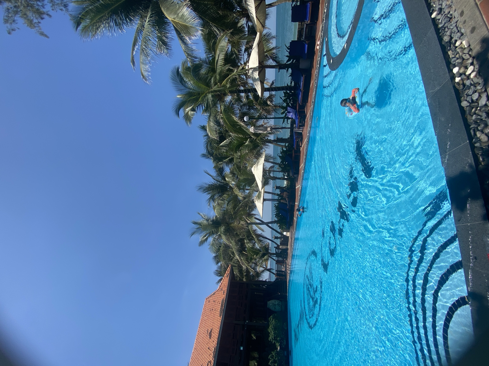 This is a full-frame, eye-level, outdoor shot of a resort pool area on a clear, sunny day, likely in Phu Thuy Ward, Vietnam, suggested by the architecture and palm trees. The foreground is dominated by a large, bright blue swimming pool. Dark, curved patterns are visible on the pool floor near the edge, and a large, dark circular logo or design is present deeper in the water.
In the pool, a single young child wearing a dark shirt and an orange flotation device is visible, appearing to be swimming. Further in the distance, two other figures are submerged in the water, with only their heads visible.
To the left of the pool, a large wooden building with an orange tiled roof is visible. Behind the building and extending across the background are numerous tall palm trees, some with coconuts, swaying slightly. Beyond the trees, the ocean is visible, with gentle waves.
Along the edge of the pool, a row of navy blue lounge chairs is set up, many with beige umbrellas providing shade. Some of these chairs appear to be occupied by two individuals. The pool deck is paved with reddish-brown tiles and is bordered by dark grey stone tiles with small, smooth grey and white pebbles. The sky is a clear, vibrant blue.