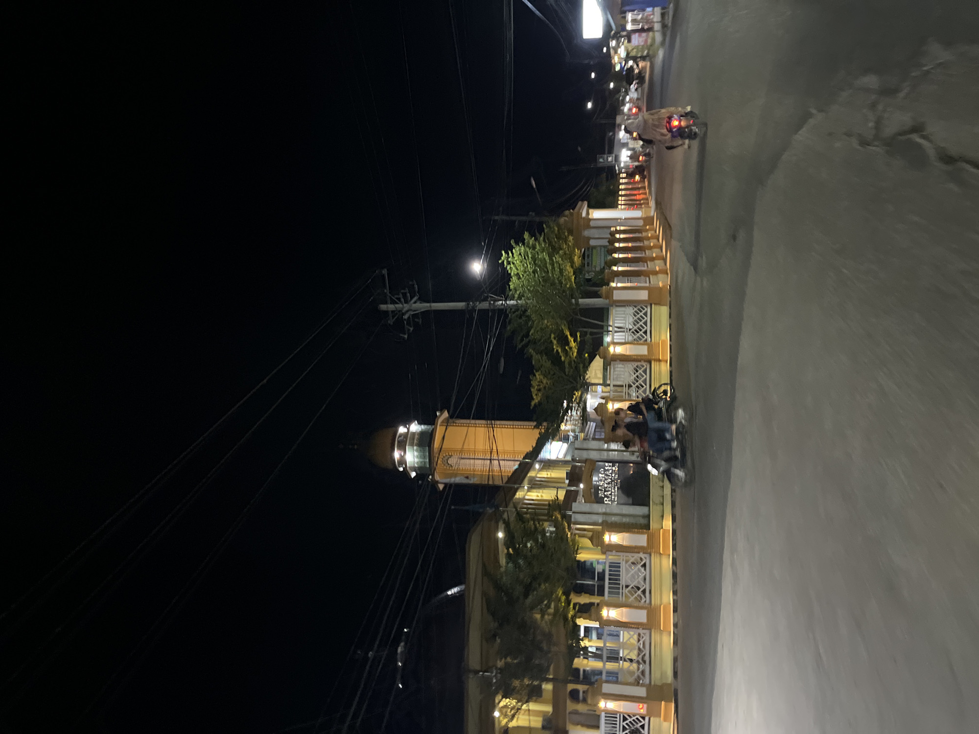 The image depicts a street scene at night in Langsa, Indonesia. A prominent yellow building with a domed minaret, identified by a sign as "MASJID RAHMAN" (Masjid Rahman), is illuminated on the left side of the frame. The building features decorative white fencing along its perimeter. In the foreground, a paved road is visible with several vehicles. On the road, a motorcycle with three passengers is moving from left to right, their figures blurred due to motion. Further down the road, another motorcycle with a single rider, wearing a headscarf and a patterned robe, is also traveling away from the camera. The sky is dark, with visible power lines and streetlights. The overall impression is a typical urban night scene with religious and transportation elements.