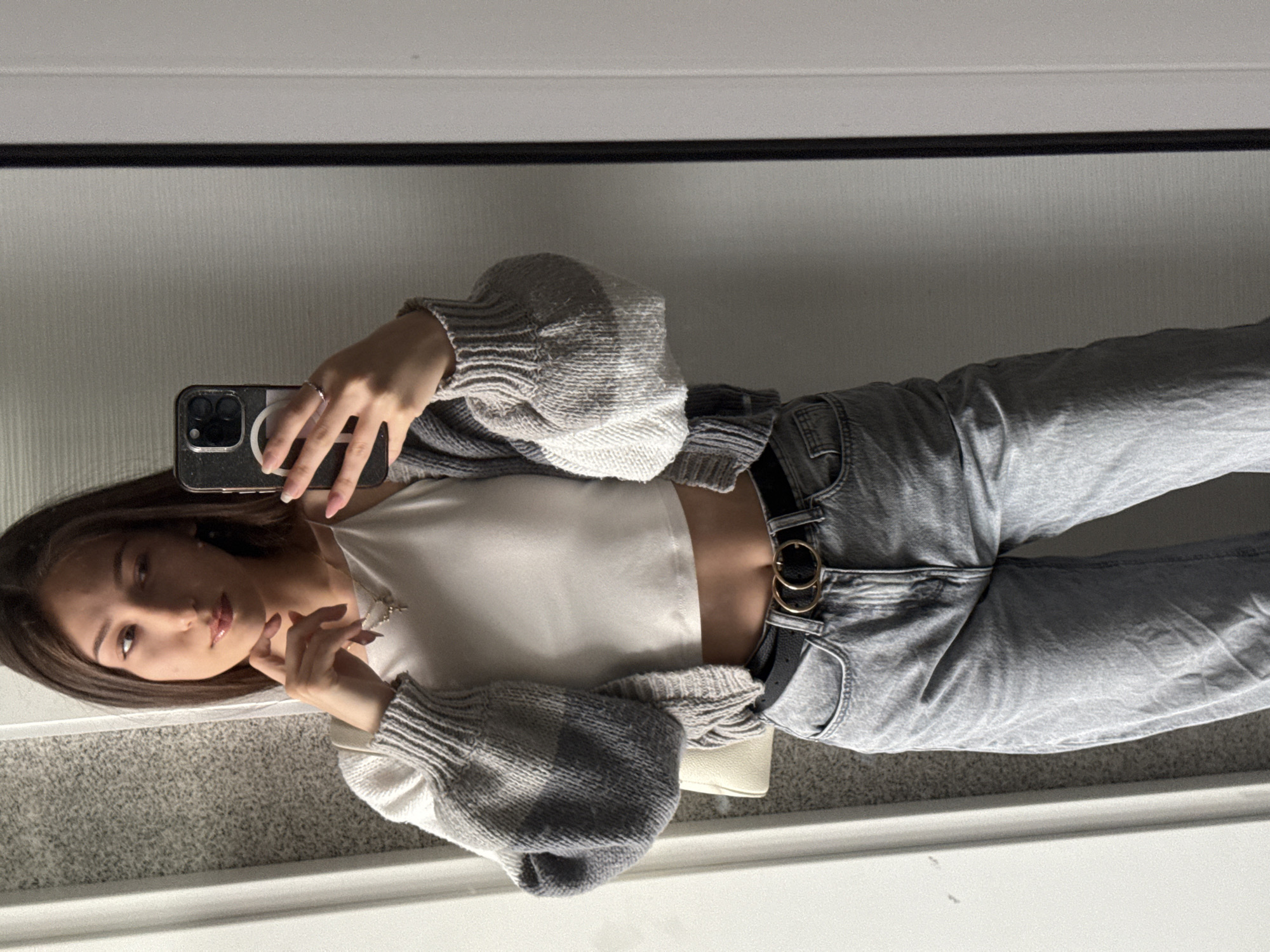 A young woman is seen in a mirror selfie. She is wearing a white crop top, a gray cardigan with voluminous sleeves, light gray jeans with a black belt featuring a double-circle gold buckle, and a silver necklace. She is holding a dark-colored smartphone in her left hand. The environment appears to be an indoor space with a reflective surface, likely a mirror. The flooring is carpeted. The image offers no discernible geographical cues indicating a location in Sofia, Bulgaria. The focus is primarily on the woman and her attire.