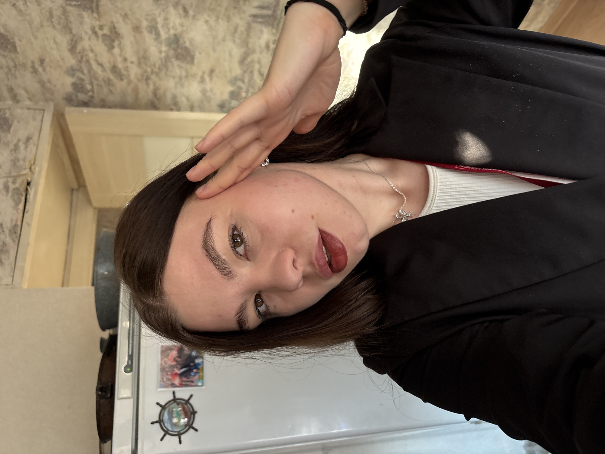 A young woman poses in front of a refrigerator. She has brown hair and green eyes. She's wearing a white top under a black blazer, and a silver necklace. Her left hand touches her forehead. The refrigerator has a magnet and a picture on it. The location is in a kitchen, with a beige counter, possibly in Perm, Russia.