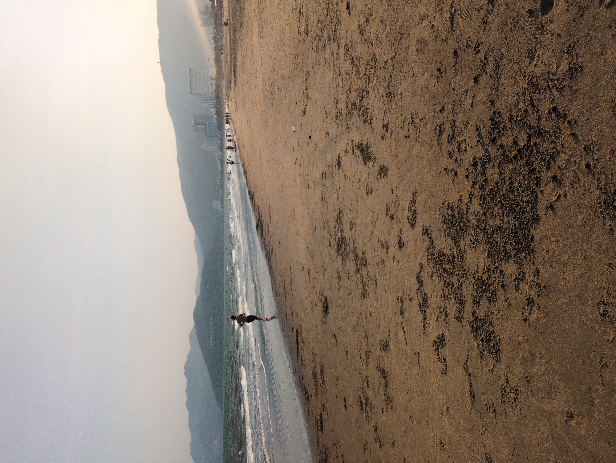 The image shows a beach scene in Quy Nhon Ward, Vietnam. A person is seen walking on the beach close to the water. The shoreline features waves lapping onto the sand. Buildings are visible along the shoreline. The sky is hazy. The beach itself is sandy and appears textured.
