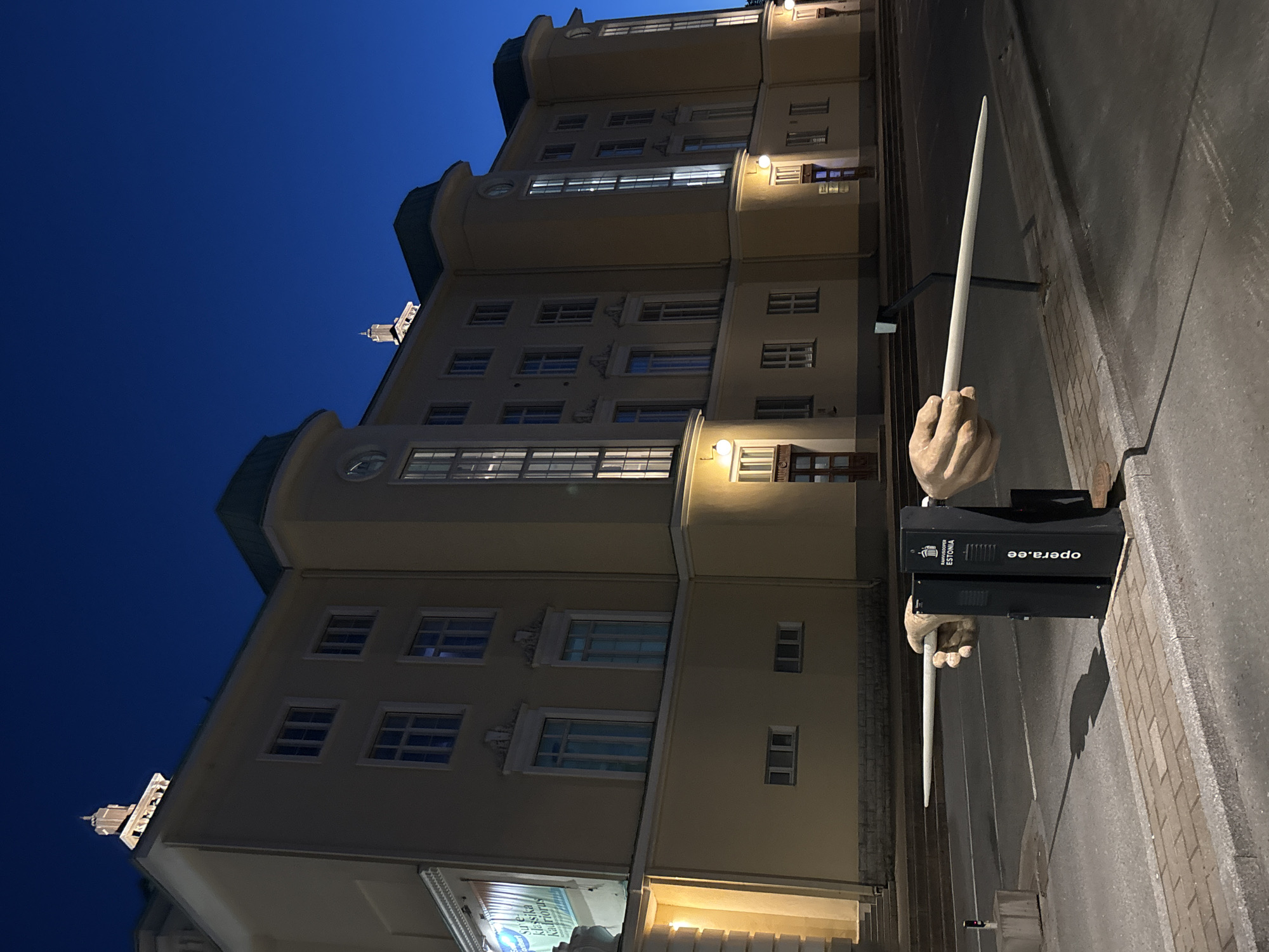 The image depicts an outdoor scene at night in Tallinn, Estonia, featuring a large, light-yellow building with multiple windows, illuminated from within and by exterior lights. In the foreground, a black kiosk with text including "opera.ee" and "RAHVOOPER ESTONIA" is prominently displayed. Two large, sculpted hands extend from the sides of this kiosk, each holding a long, white, baton-like object horizontally. The hands and batons are part of an art installation. The building appears to be a public institution, possibly an opera house or cultural center, given the text on the kiosk. The sky is a deep, dark blue, indicating nighttime. The ground in the foreground consists of a paved sidewalk and an asphalt road.