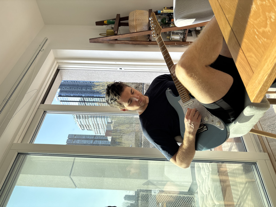 A male individual, appearing approximately 30-40 years old with short dark hair, is seated on a light grey upholstered chair, actively playing a light blue Ibanez electric guitar. He wears a dark short-sleeved t-shirt and dark shorts, with his left leg crossed over his right, supporting the guitar. His hands are engaged with the fretboard and strings. The setting is an indoor residential space, brightly illuminated by natural sunlight entering through a large, floor-to-ceiling window unit on the left. Outside the window, a modern urban environment is visible under a clear blue sky, featuring multiple high-rise buildings, prominently including the distinctively curved and layered facade of the Lexicon building (270 City Road), situating the location in Islington, London, United Kingdom. To the right of the window, a multi-tiered wooden ladder-style shelf unit holds various bottles and a woven basket. A light wooden table is partially visible in the foreground to the right.