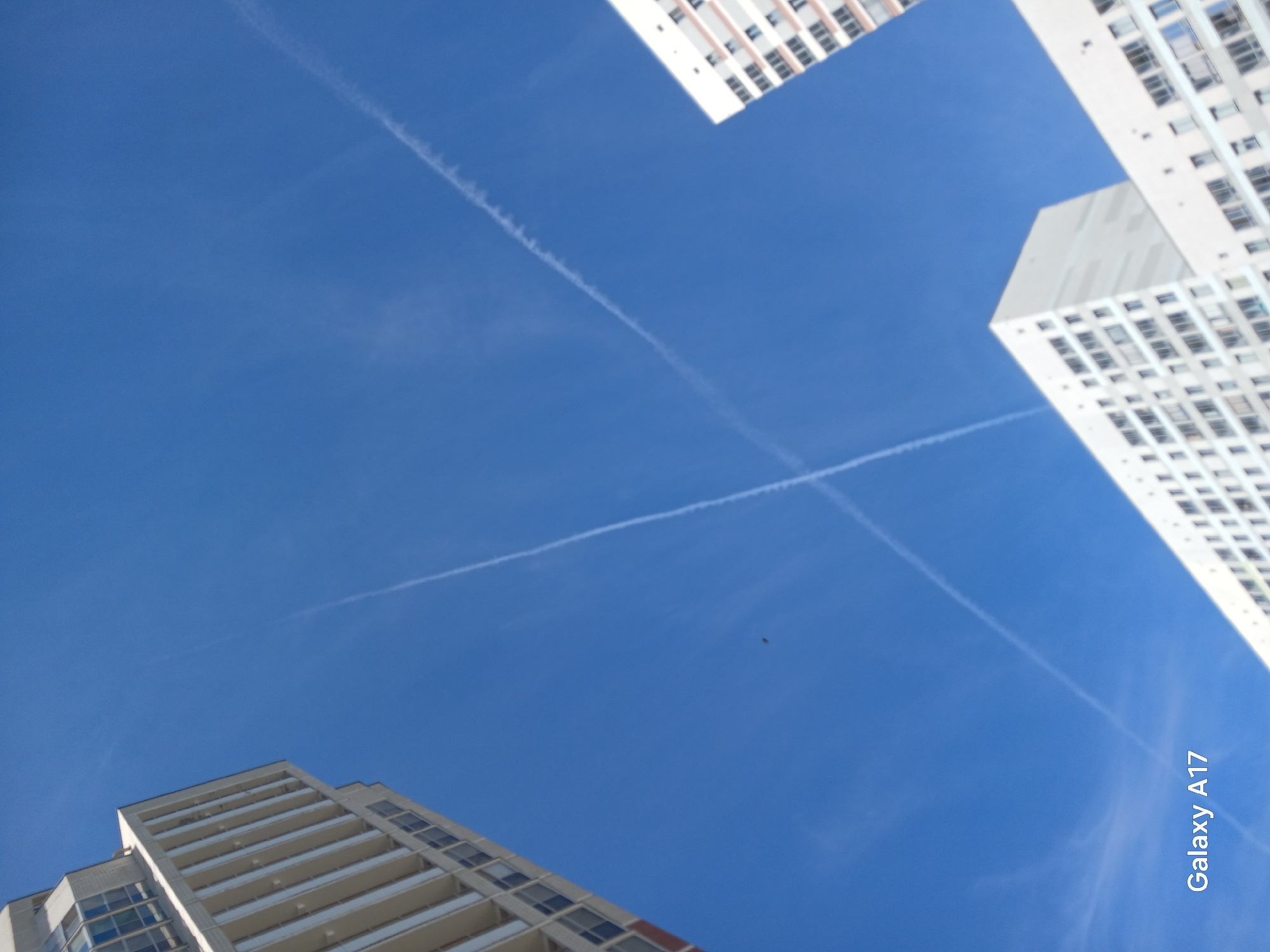 The image captures a view upwards towards a clear, blue sky, framed by three tall buildings. The buildings are white with numerous windows, suggesting residential or office complexes. The camera perspective is tilted upwards, giving a distorted view of the buildings. Vapor trails from airplanes form a cross in the sky, adding a dynamic element. A small, dark object, possibly a bird, is visible in the upper-center of the frame. The text "Galaxy A17" is visible in the lower-right corner, likely indicating the device used to capture the image. The location is the city of Yekaterinburg, Russia.