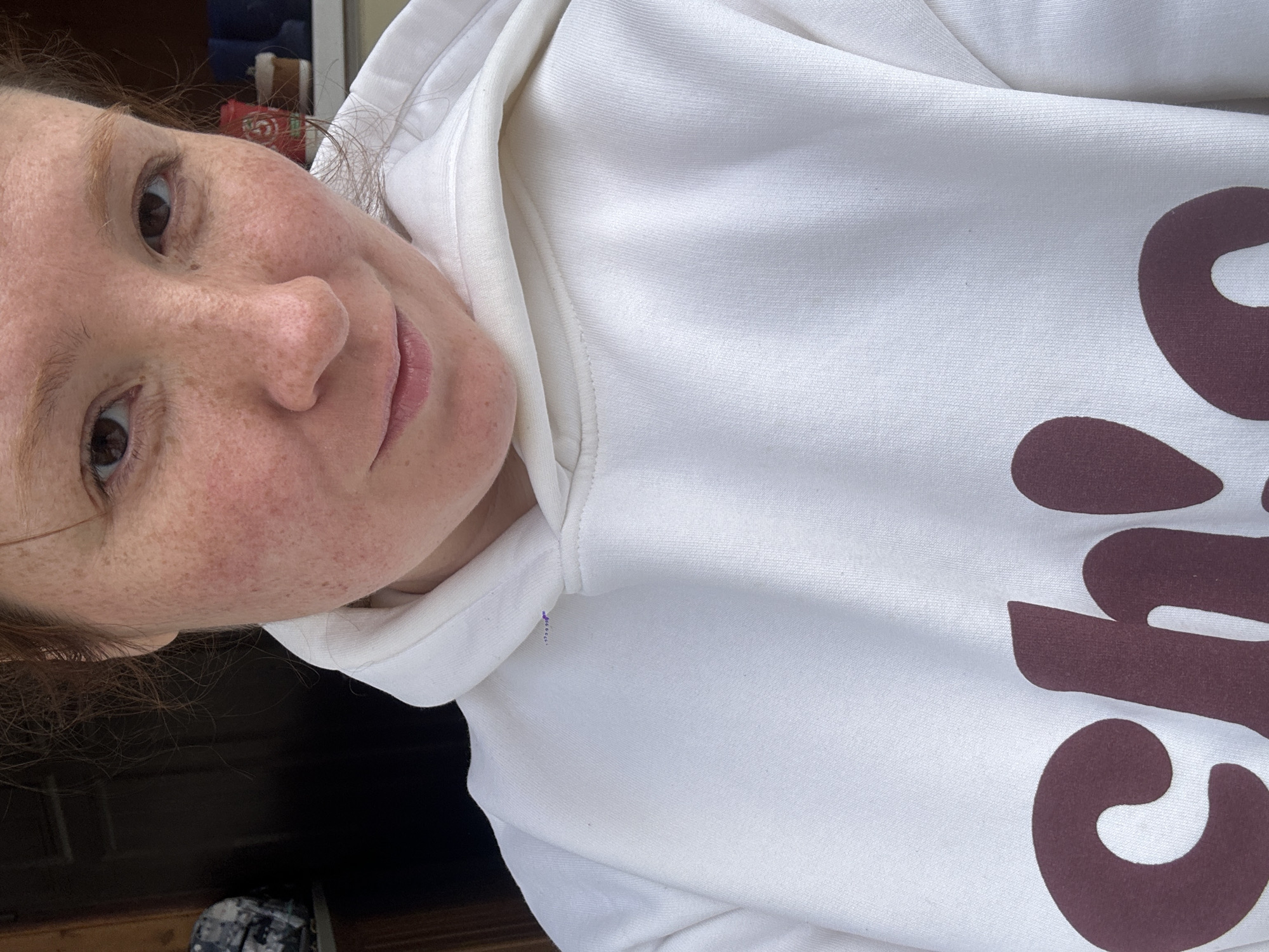 A close-up photograph shows a person with fair skin and freckles wearing a white hooded sweatshirt with brown lettering. The person is positioned at an angle, with their face visible and part of the sweatshirt's design showing. The background is partially visible and appears to be an interior setting with dark-colored elements. There is no evidence suggesting that the photo was taken in Nizhny Novgorod, Russia.