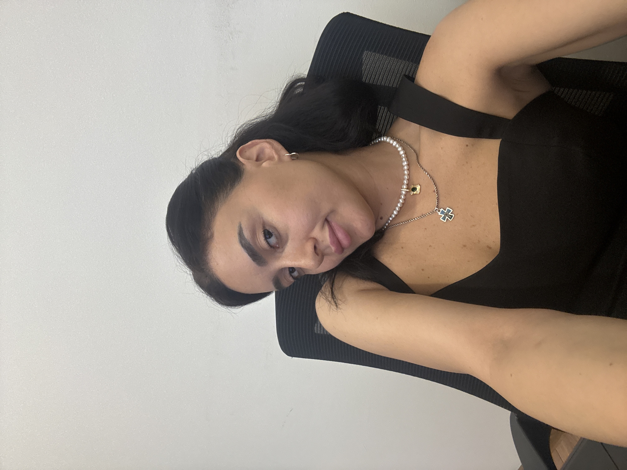 A woman with dark hair and light skin is sitting in a black office chair against a plain white wall. She is wearing a black dress and several necklaces, including one with pearls and a cross pendant, and another with a small gold charm. She is looking towards the camera with her head tilted slightly. Her left arm is extended forward, resting on the chair. The image appears to be a selfie taken indoors, likely in an office setting. There is no indication of the city of Tbilisi, Georgia.