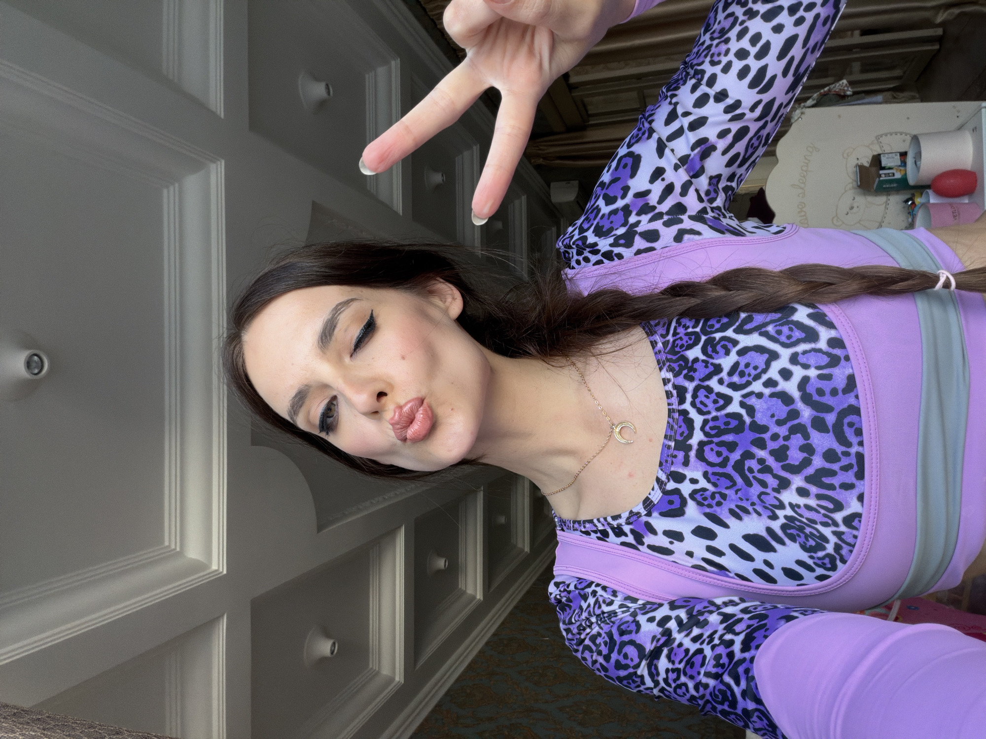 A young woman with dark hair in a braid, wearing a purple leopard print long-sleeved shirt and a lavender sports bra, winks and makes a peace sign with her right hand. She is pouting her lips and has dramatic winged eyeliner. A crescent moon pendant hangs from a delicate gold necklace around her neck. She is indoors, with a detailed white ceiling featuring recessed lighting and ornate molding visible above her. The background shows patterned wallpaper, curtains, and a child's crib with stuffed animals. The image appears to be a selfie taken with a smartphone. There is no visible indication of the city of Ufa, Russia.